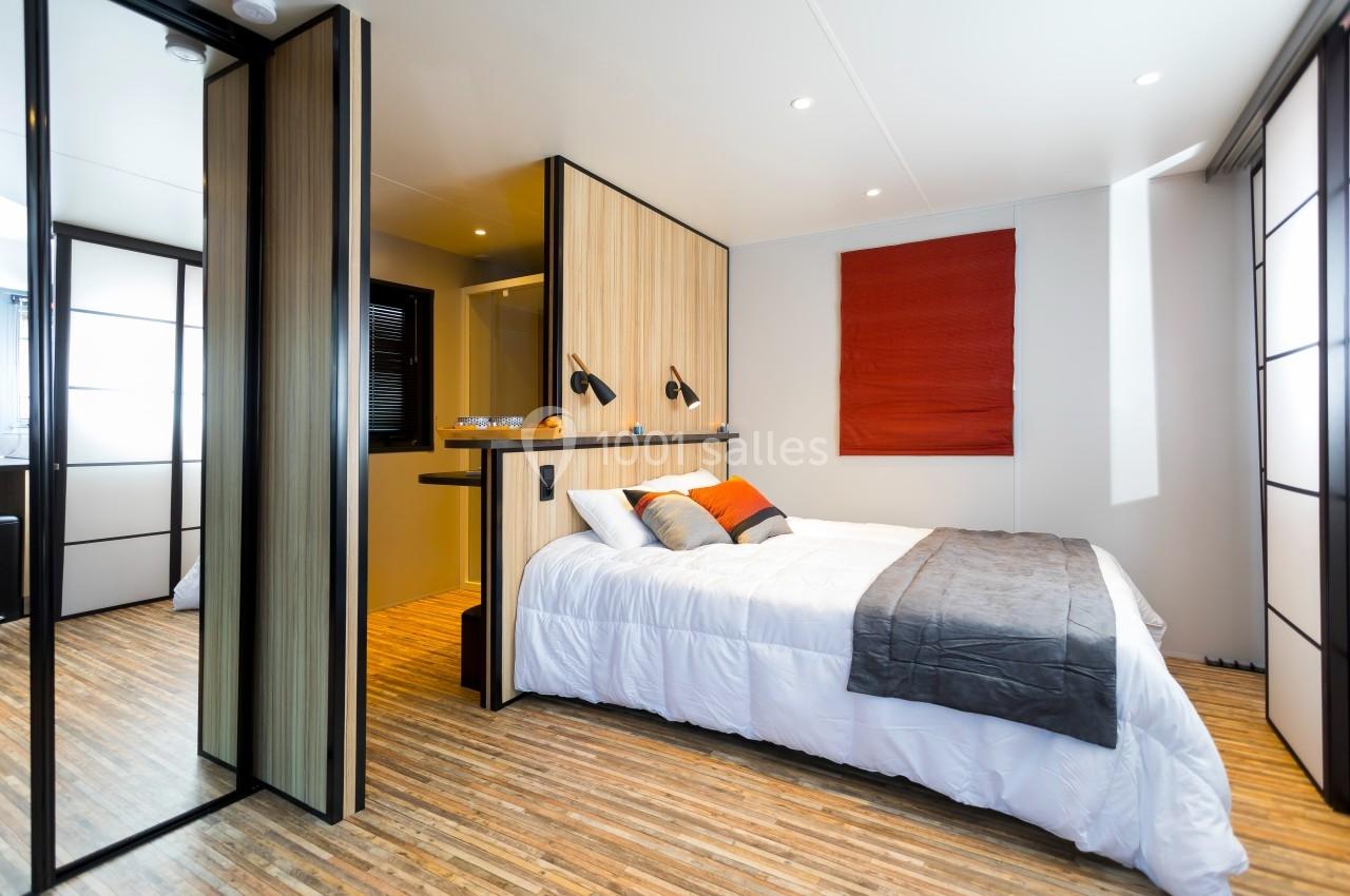 Chambre moderne avec lit double, parquet clair, cloison en bois, éclairage mural et store rouge.
