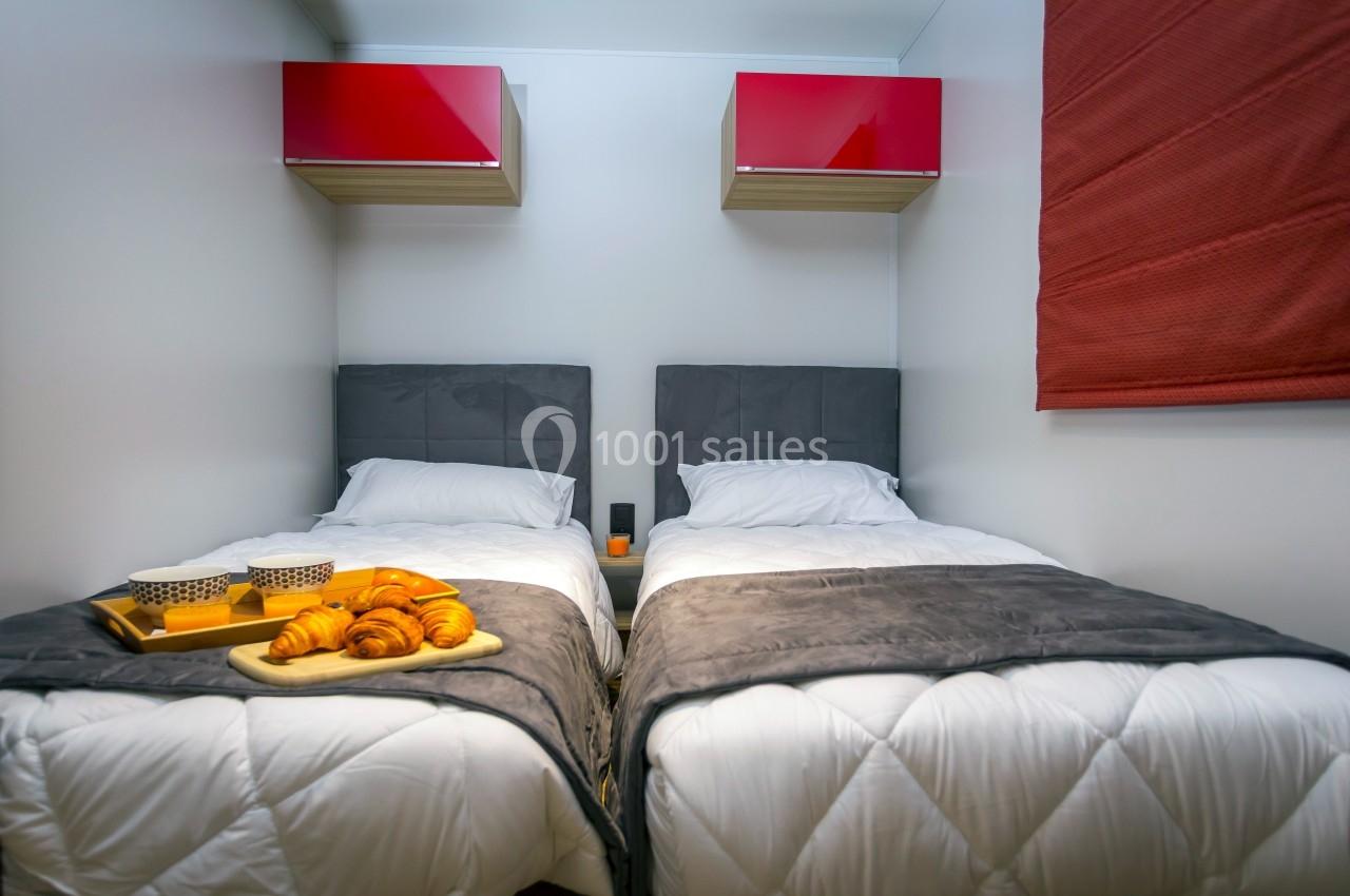 Deux lits simples avec couettes blanches, plateau de petit-déjeuner posé sur l'un, étagères rouges au mur.