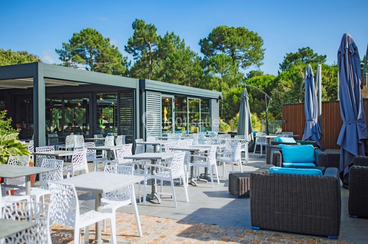 Terrasse extérieure avec tables, chaises blanches et fauteuils en rotin, entourée de verdure sous un ciel bleu.