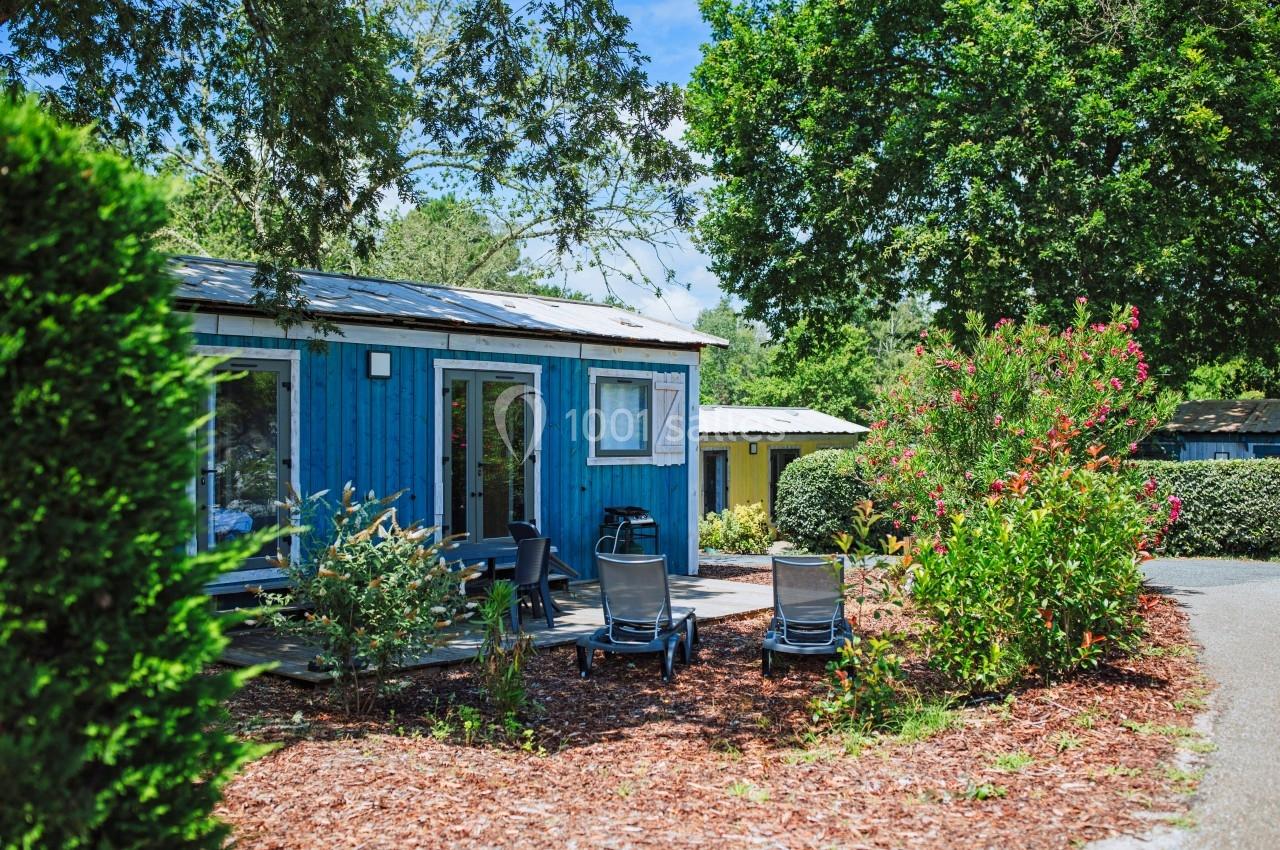 Chalet en bois bleu entouré de végétation, avec une terrasse aménagée et des chaises sous un ciel ensoleillé.