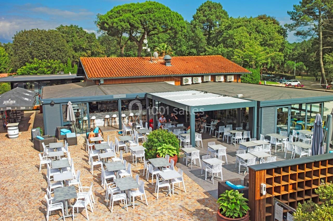 Terrasse d'un restaurant avec tables et chaises en extérieur, entourée de verdure et d'un bâtiment à toit rouge.