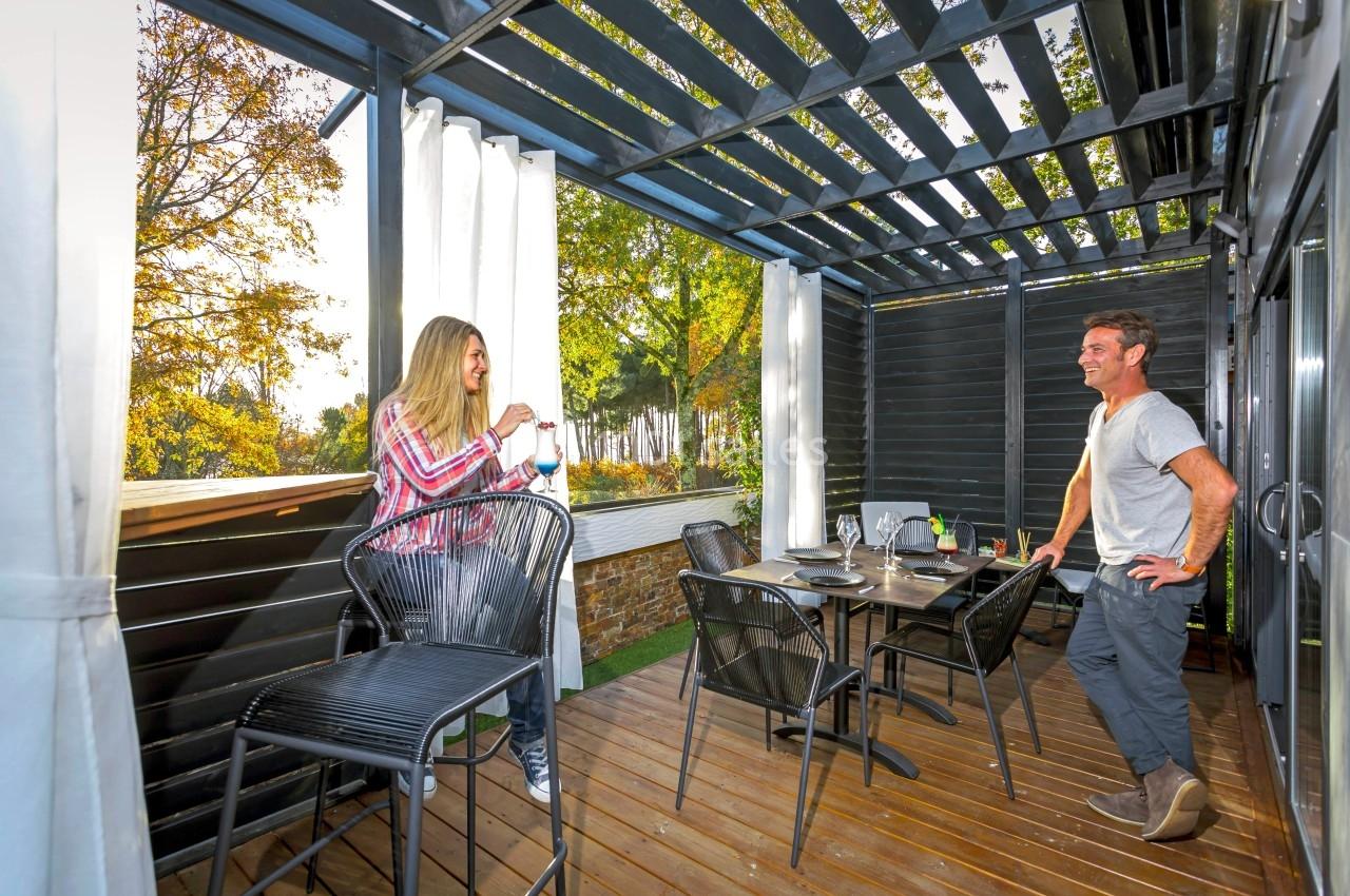 Une femme et un homme discutent sur une terrasse en bois aménagée avec une table dressée et des chaises hautes.