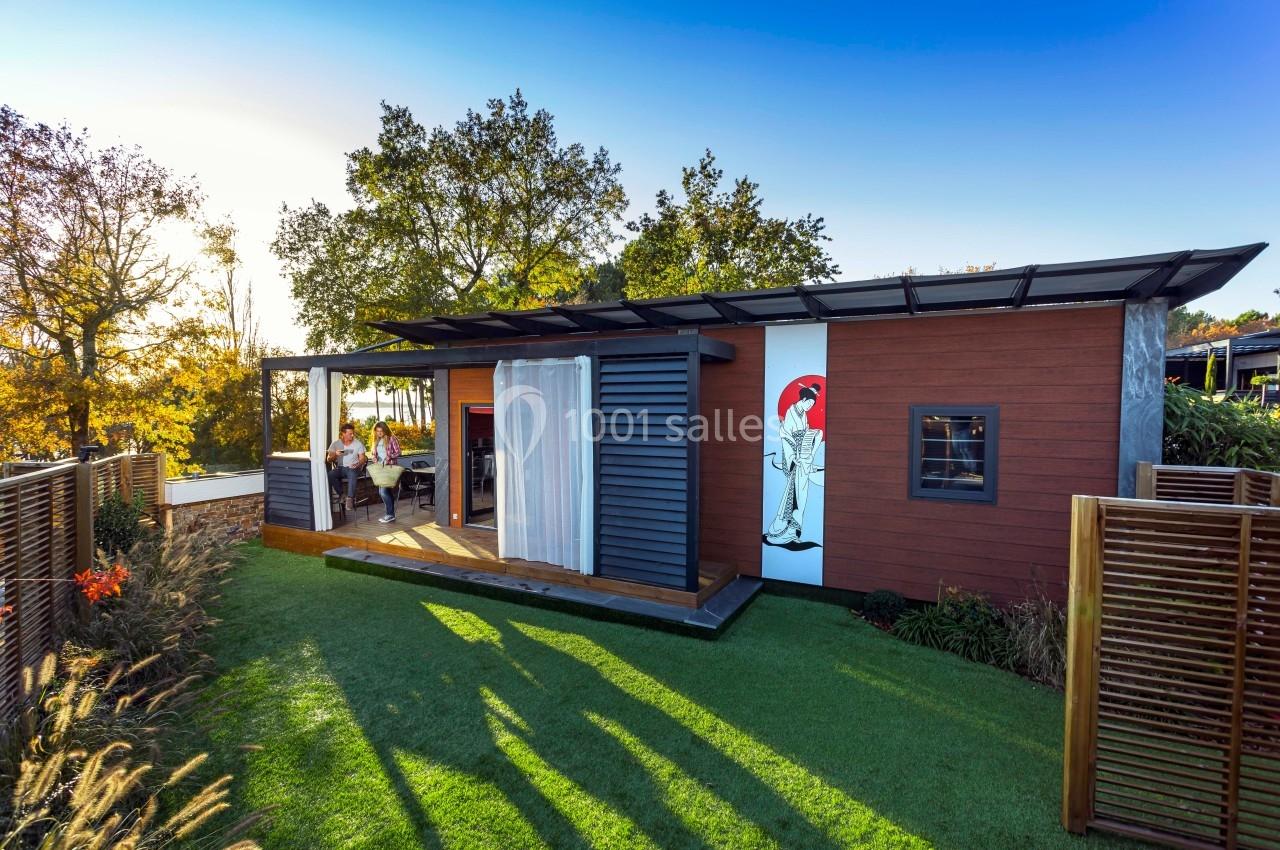 Maison modulaire en bois avec terrasse, située dans un jardin verdoyant sous un ciel ensoleillé.