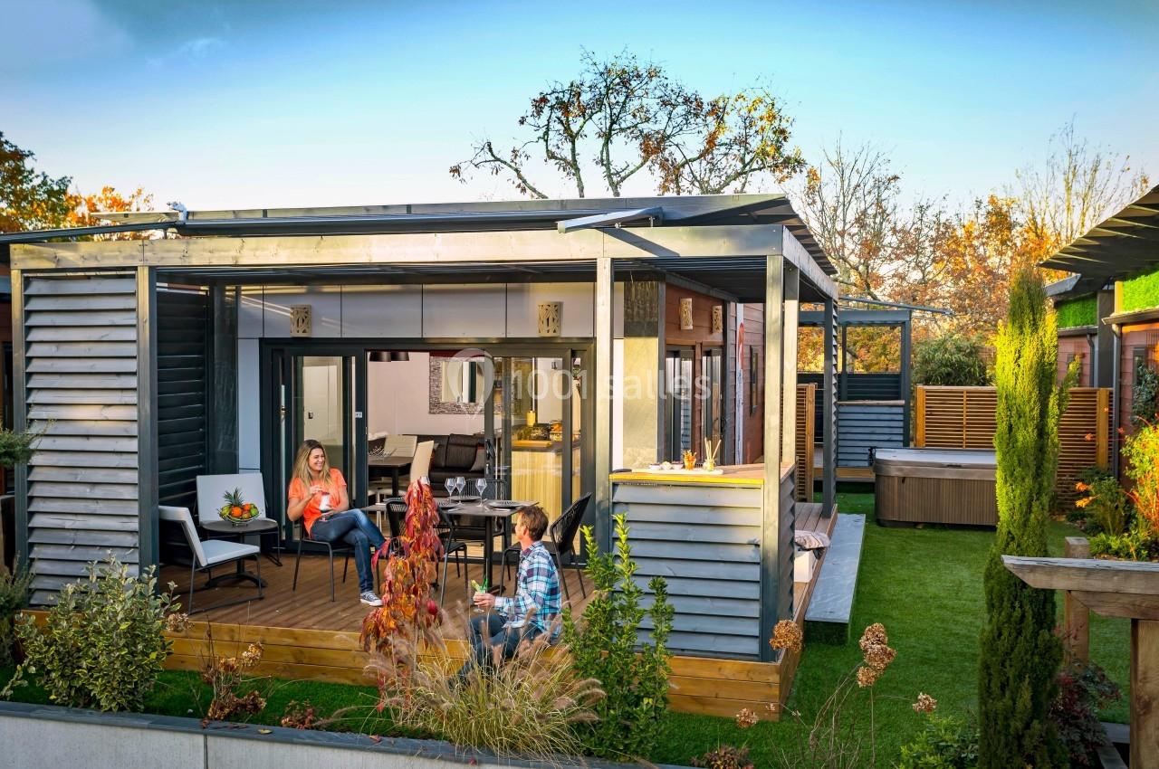 Deux personnes discutent sur la terrasse en bois d'une maison moderne entourée de végétation.