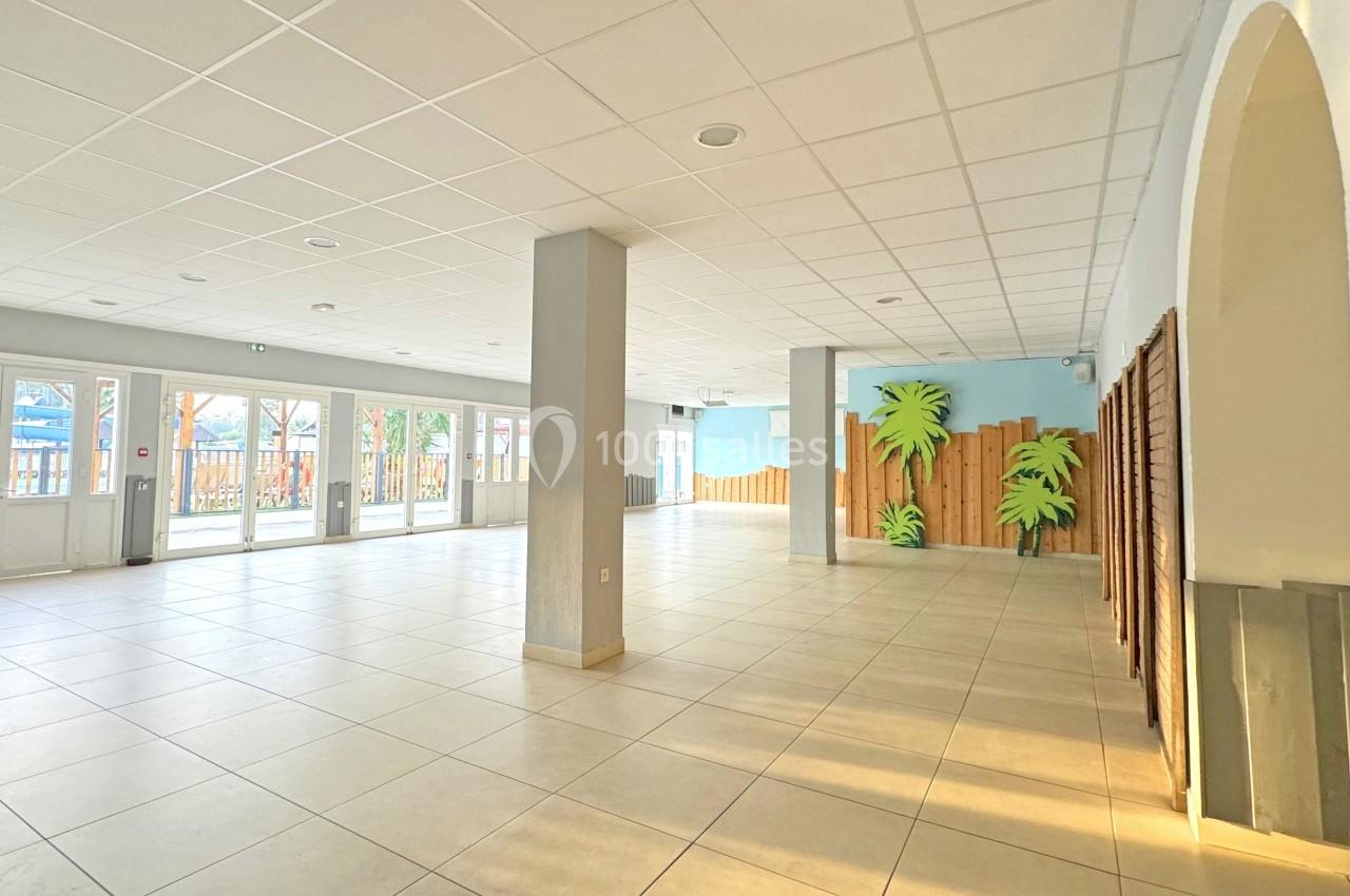 Salle lumineuse avec carrelage clair, grandes baies vitrées, piliers centraux et décorations murales de palmiers.