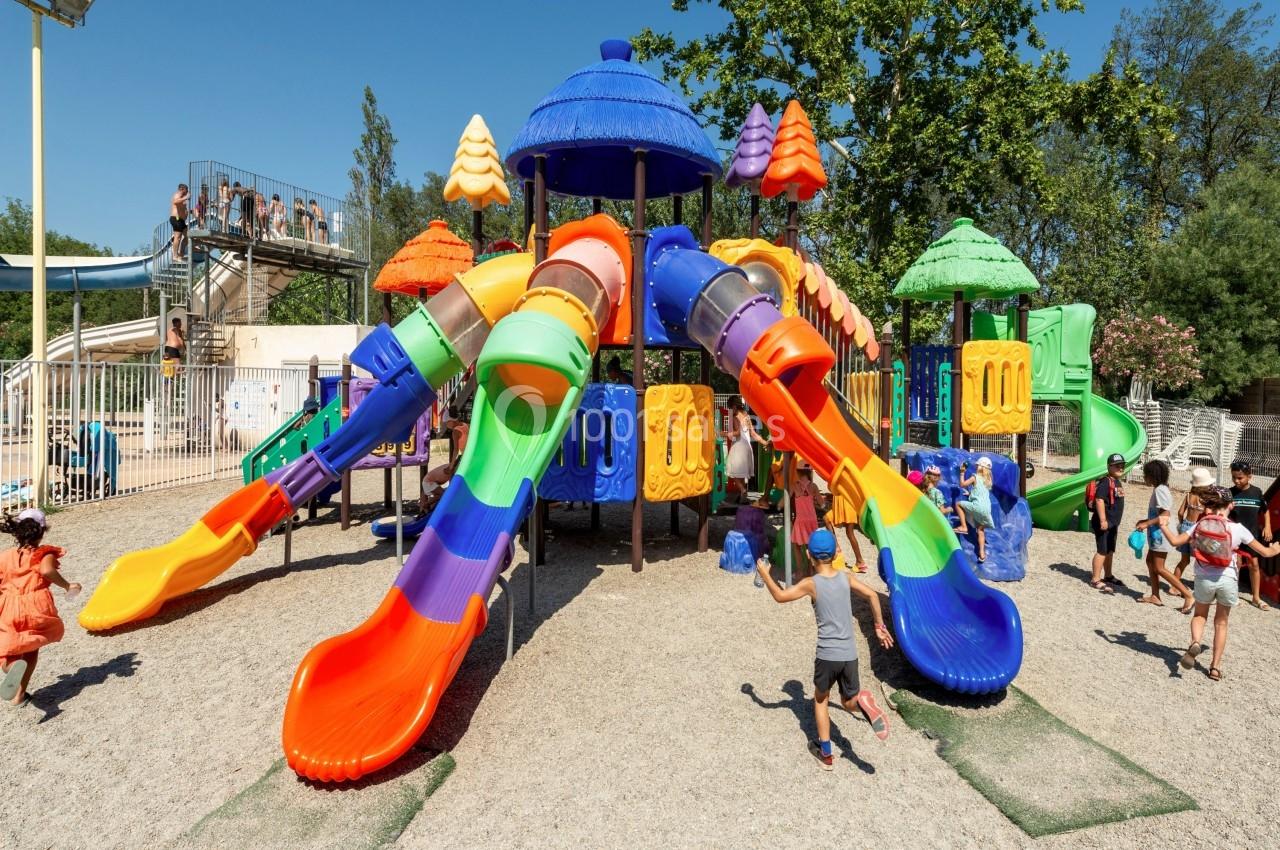 Structure de jeux colorée avec toboggans multiples, entourée d'enfants jouant sur un sol en gravier, par temps ensoleillé.