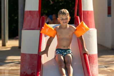 Un enfant avec des brassards orange glisse sur un toboggan rouge dans une piscine extérieure ensoleillée.