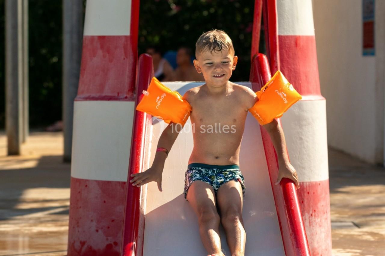 Un enfant avec des brassards orange glisse sur un toboggan rouge dans une piscine extérieure ensoleillée.