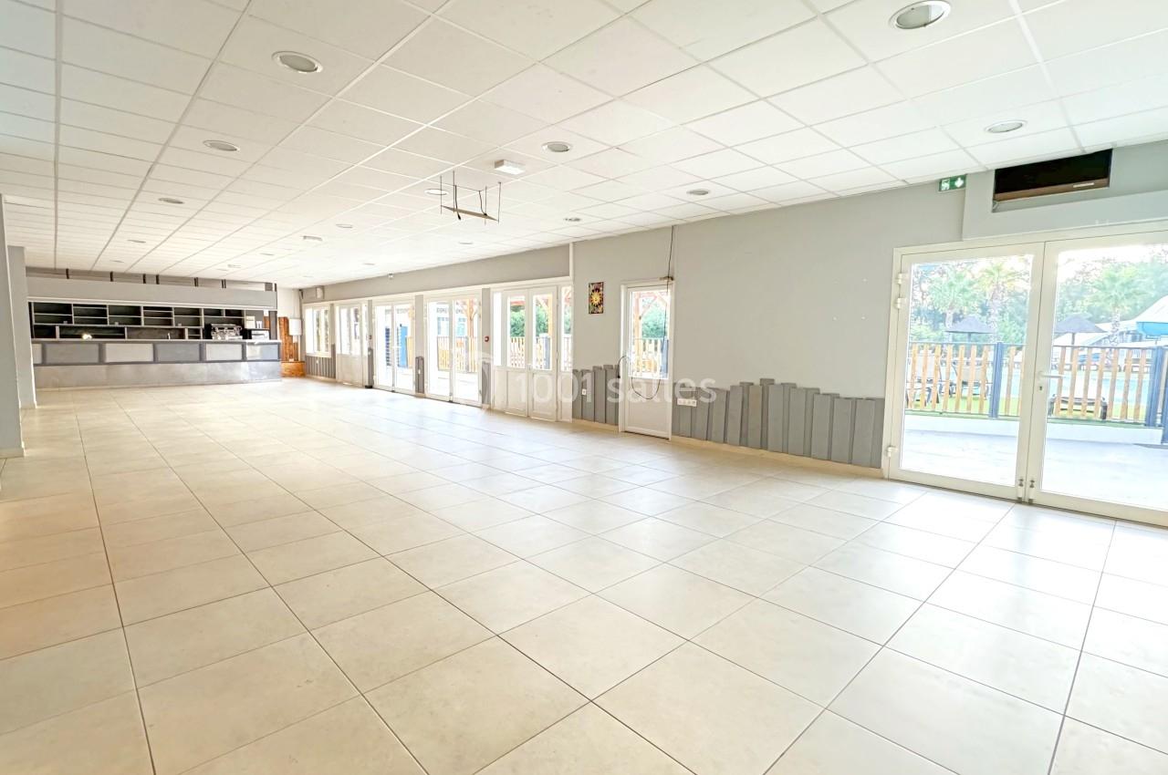 Salle lumineuse avec carrelage clair, grandes baies vitrées, comptoir à l’arrière et vue sur un espace extérieur.