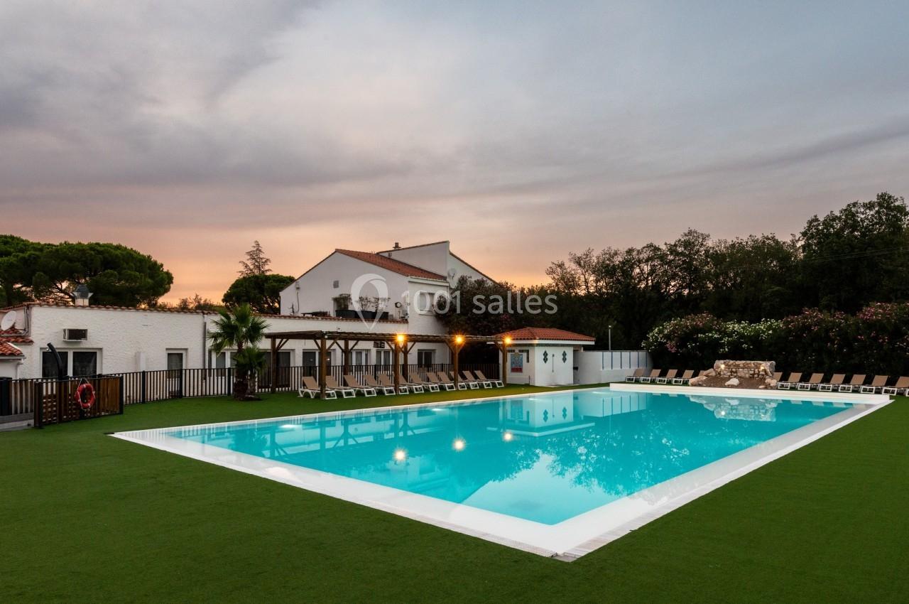 Piscine extérieure éclairée entourée de chaises longues, pelouse artificielle et bâtiments blancs au crépuscule.