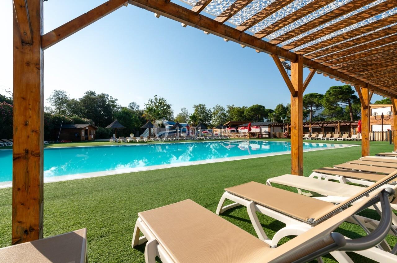 Piscine extérieure entourée de chaises longues sous une pergola en bois, avec des arbres en arrière-plan.