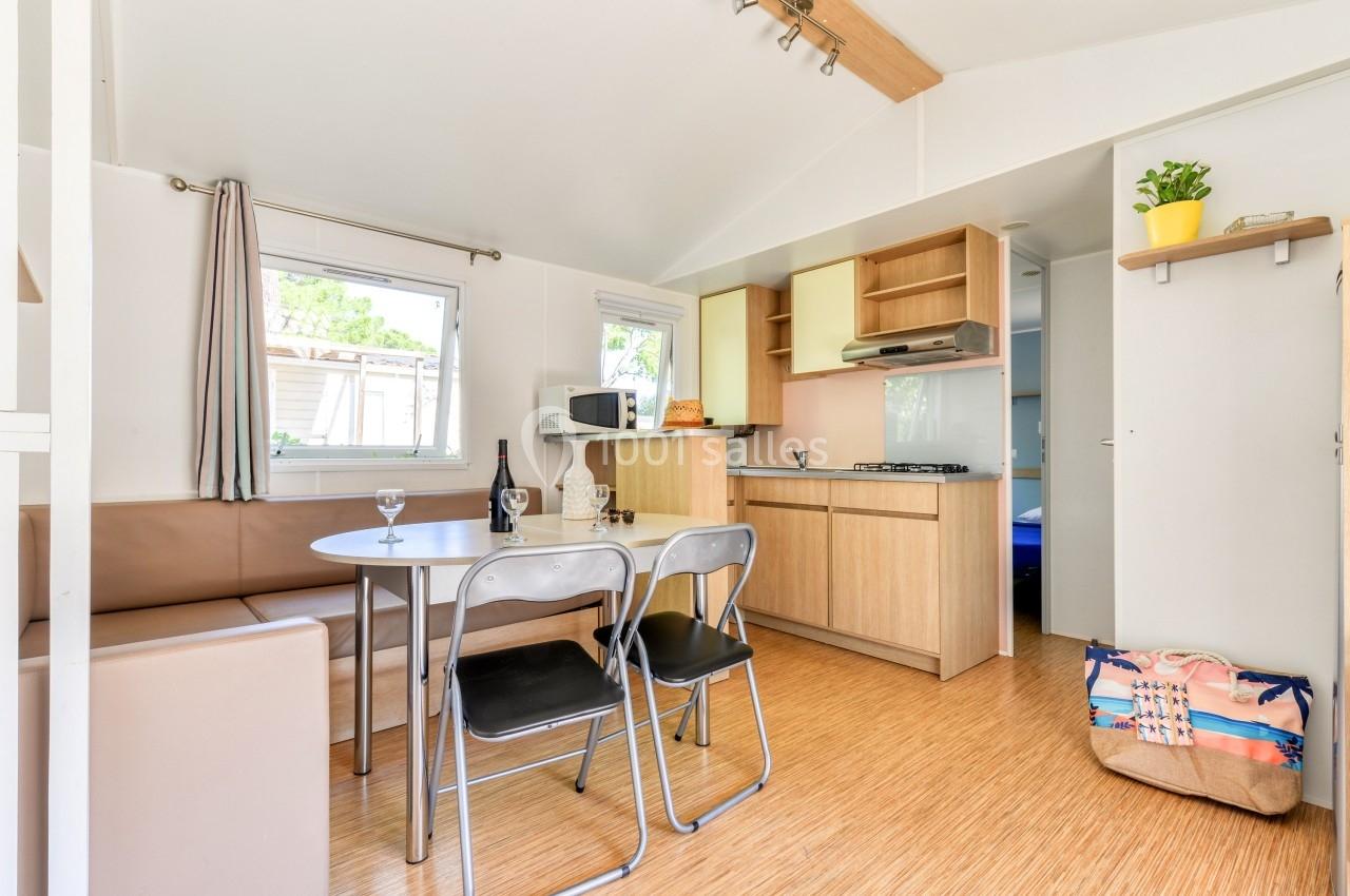 Intérieur d'un mobil-home avec coin salon, cuisine équipée, table, chaises et sac posé au sol.