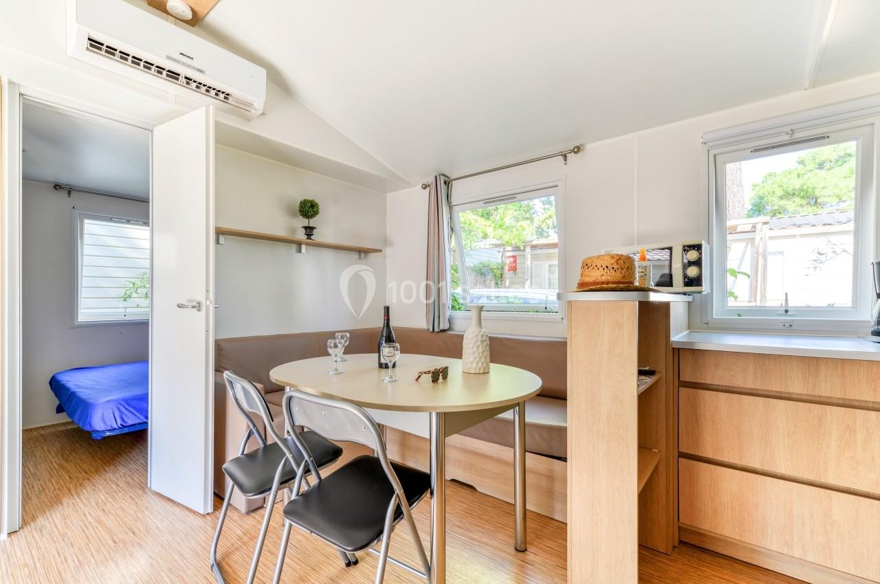 Intérieur lumineux d'un mobil-home avec table, chaises, coin cuisine et vue sur une chambre à l'arrière.