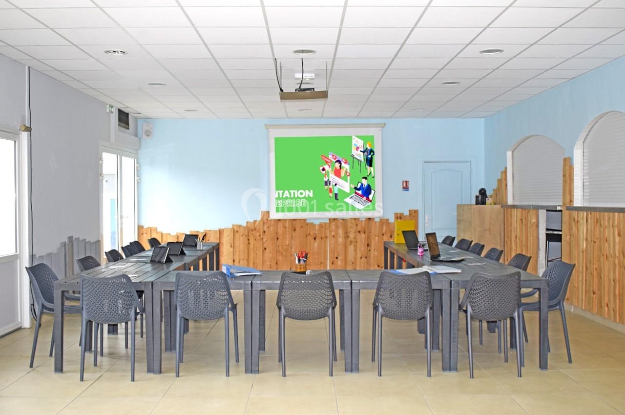 Salle de réunion lumineuse avec tables disposées en U, chaises grises et écran vert affichant une présentation.