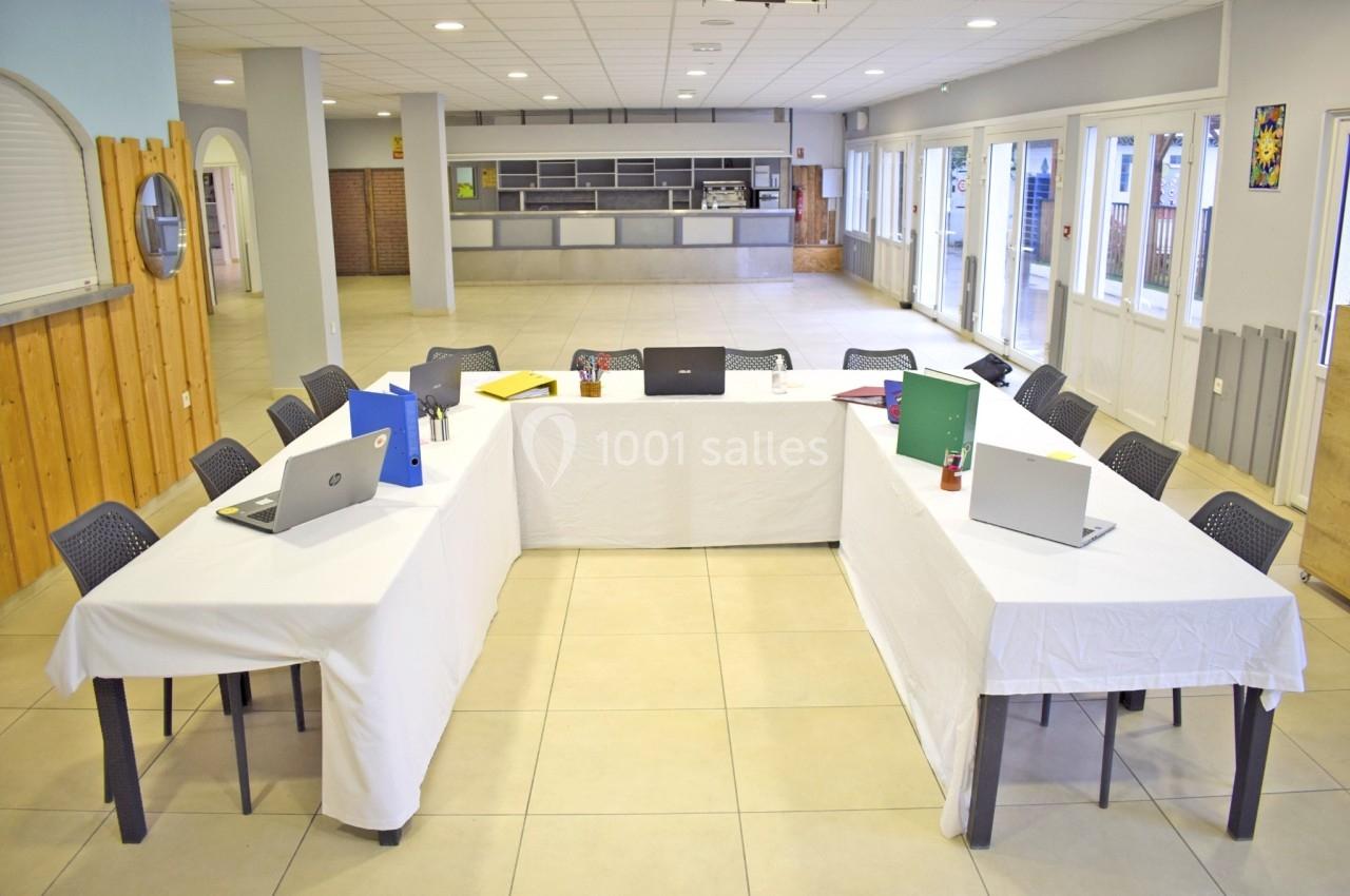 Salle lumineuse avec tables en U couvertes de nappes blanches, ordinateurs portables et dossiers colorés posés dessus.