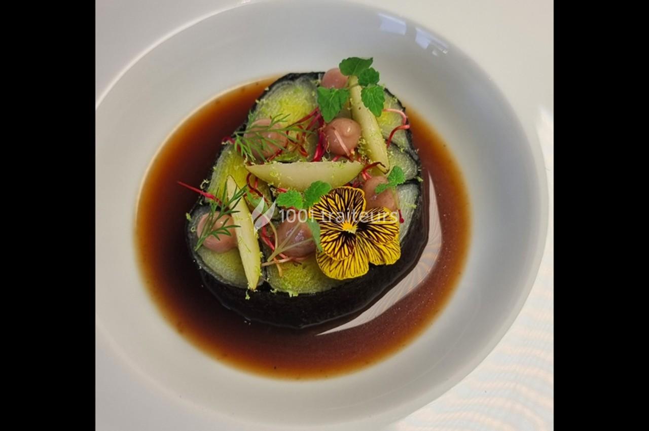 Assiette gastronomique avec un plat végétal coloré, décoré de fleurs comestibles et servi dans un bouillon brun.