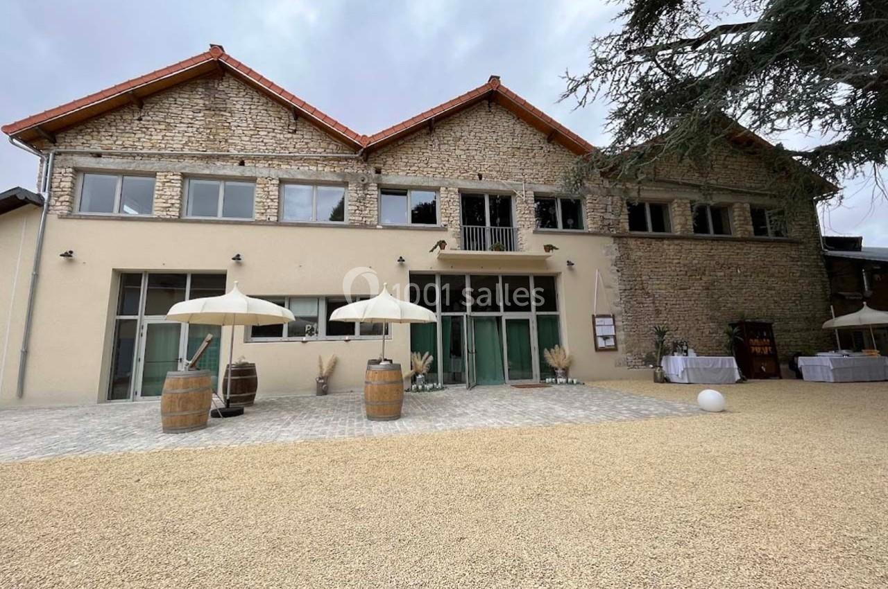 Façade d'un bâtiment en pierre rénové avec grandes baies vitrées, parasols et tonneaux sur une cour gravillonnée.