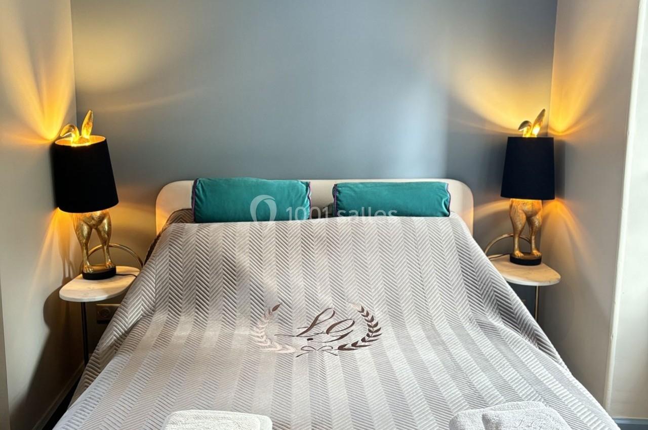 Lit double avec couverture grise, deux coussins turquoise, et lampes de chevet allumées sur des tables en bois.