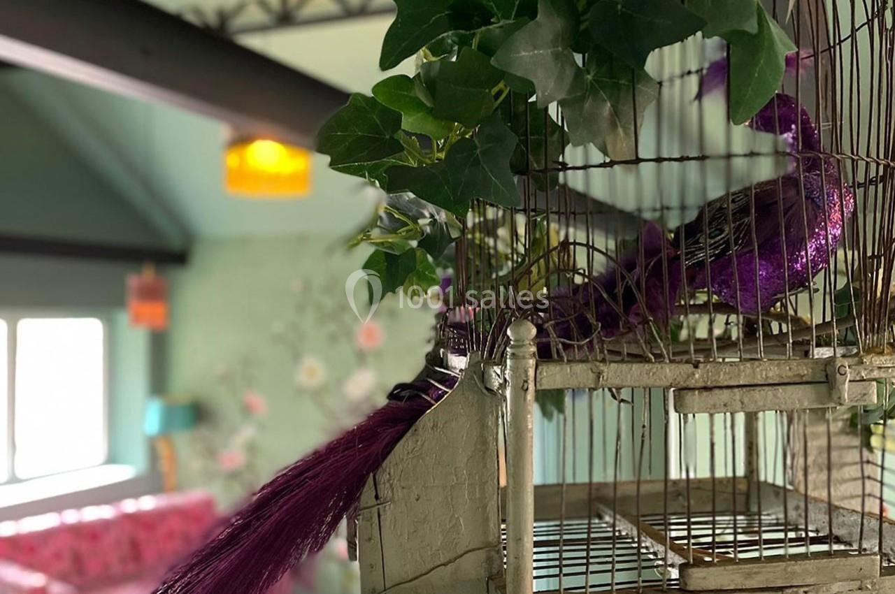 Oiseau décoratif violet avec une longue queue dans une cage en métal, entouré de feuillage, dans un intérieur lumineux.