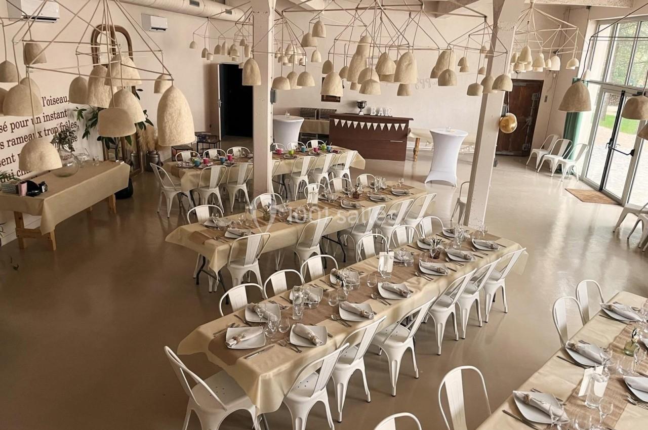 Salle de réception lumineuse avec tables dressées, chaises blanches et décorations suspendues en fibres naturelles.