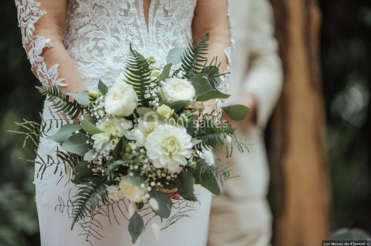 Robe de mariée en dentelle avec un bouquet de fleurs blanches et feuillage vert, tenue dans un cadre naturel.