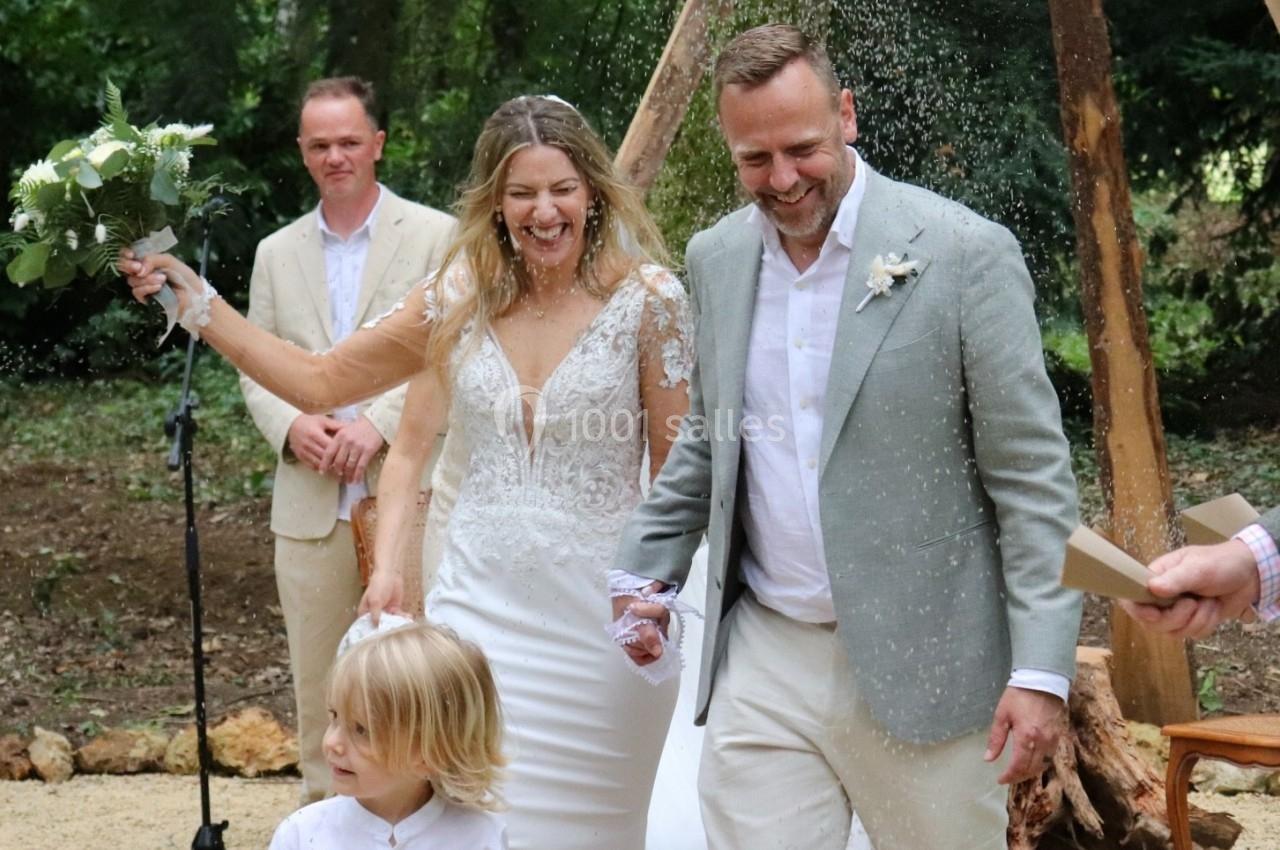 Un couple souriant sort d'une cérémonie de mariage en plein air, accompagné d'un enfant et entouré de confettis.