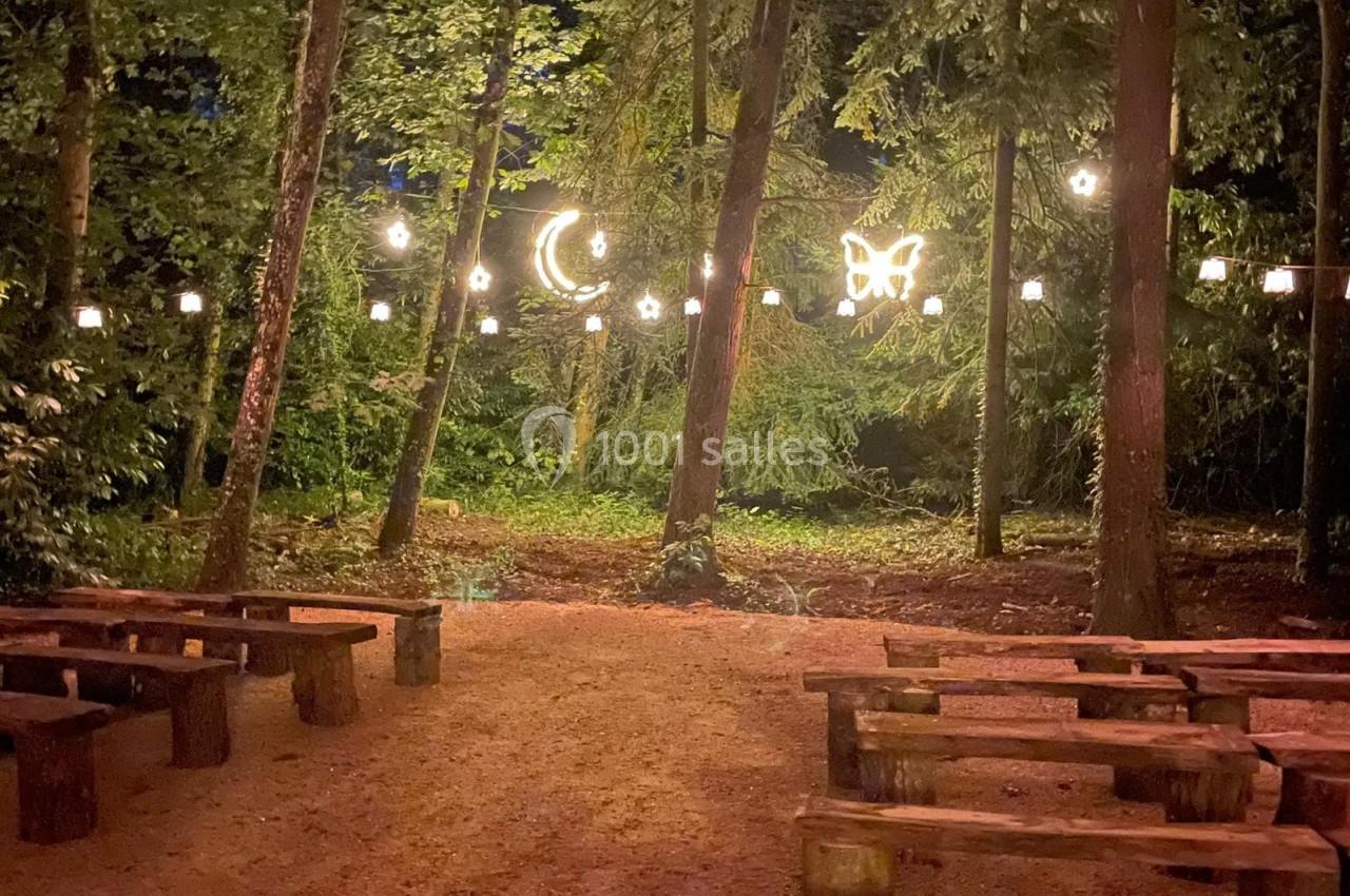 Bancs en bois disposés en extérieur sous des guirlandes lumineuses avec des motifs de lune et papillon dans une forêt.