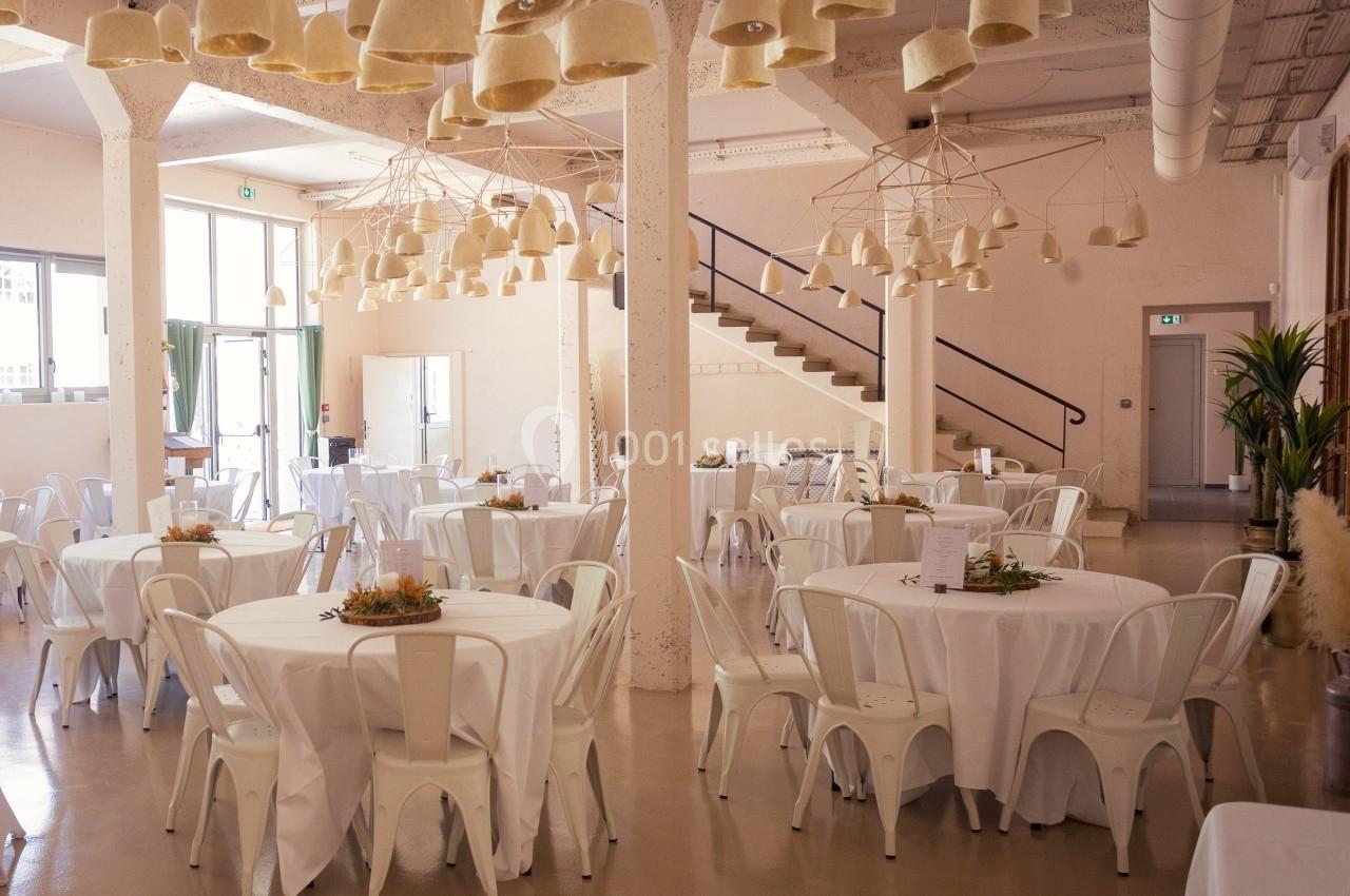 Salle lumineuse avec tables rondes dressées de nappes blanches, chaises blanches et suspensions en fibre naturelle.