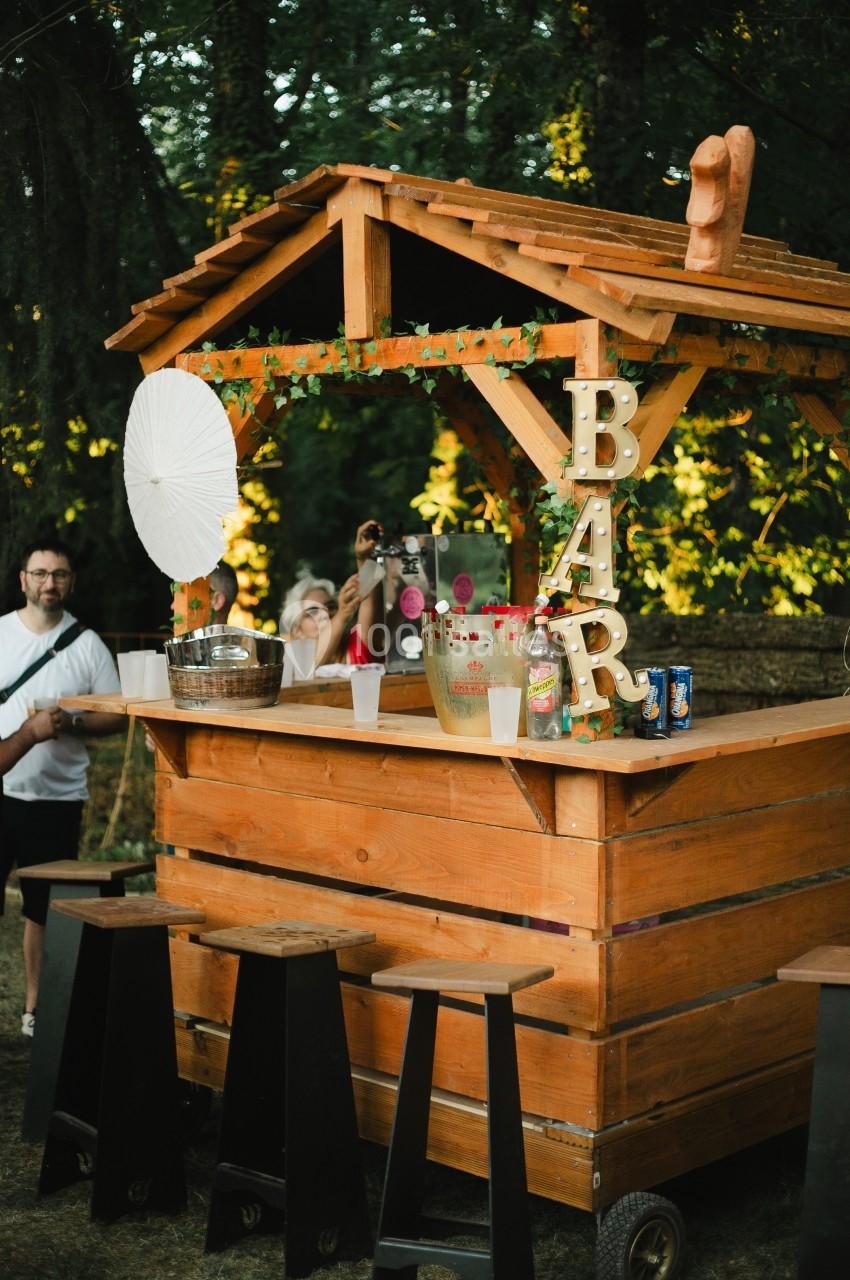 Bar en bois extérieur avec tabourets noirs, décoré de lumières et feuilles, entouré de personnes dans un cadre boisé.