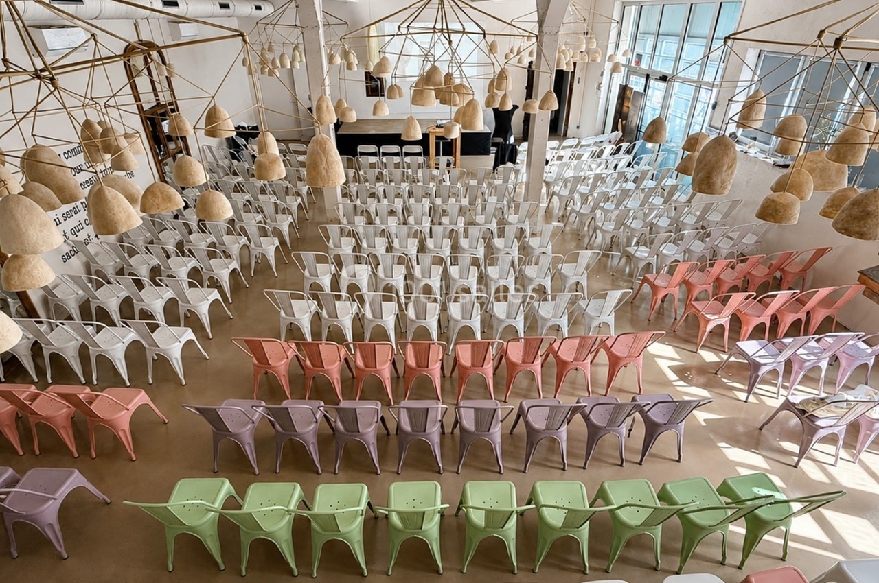 Salle lumineuse avec des rangées de chaises colorées en métal, disposées face à une scène avec un comptoir en arrière-plan.
