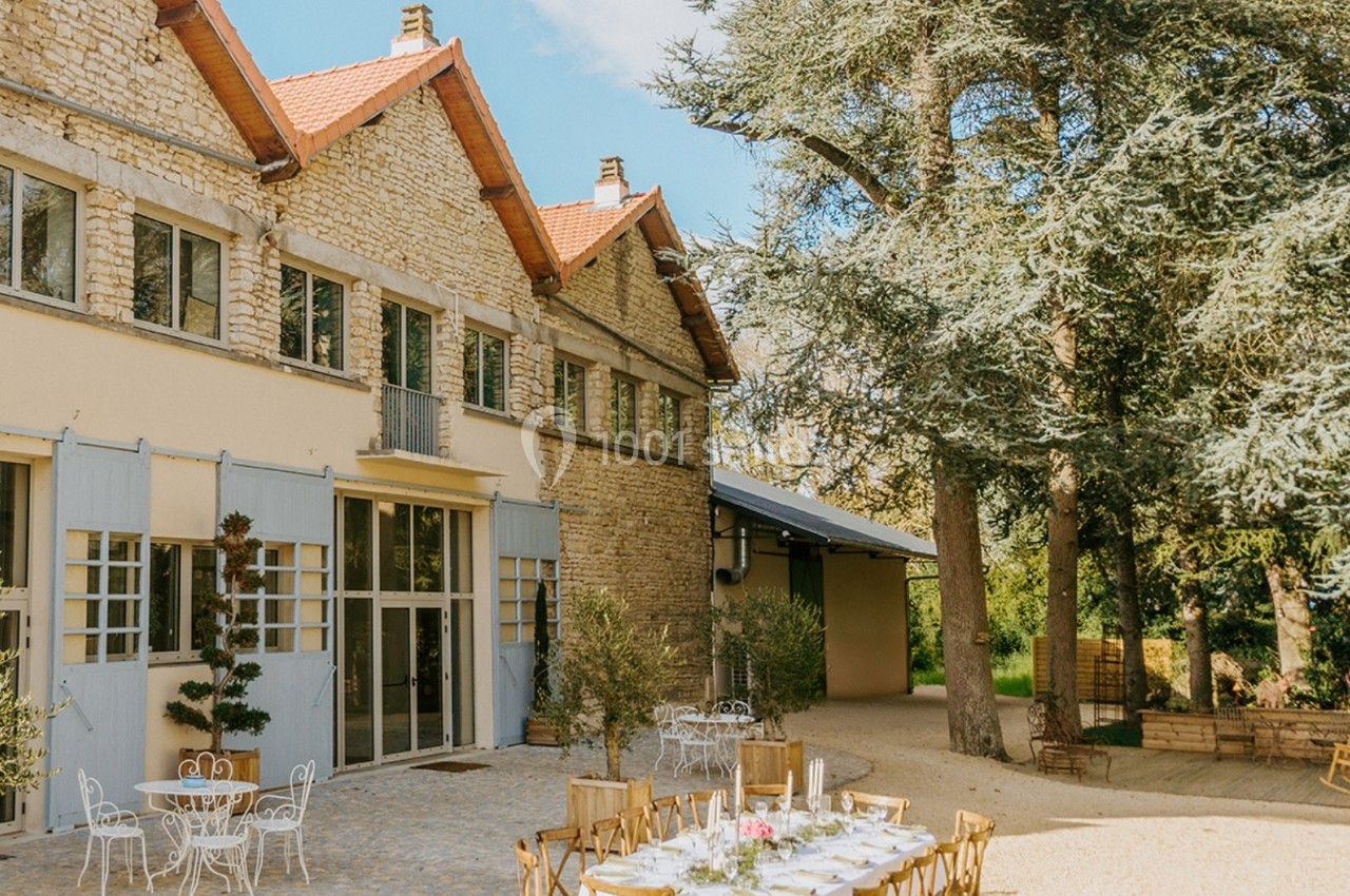 Cour extérieure d'une maison en pierre avec tables et chaises installées sous des arbres, par temps ensoleillé.
