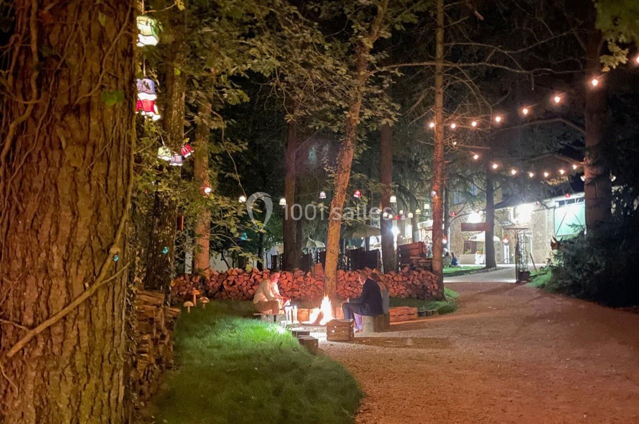 Des personnes assises autour d'un feu de camp dans une clairière boisée éclairée par des guirlandes lumineuses.