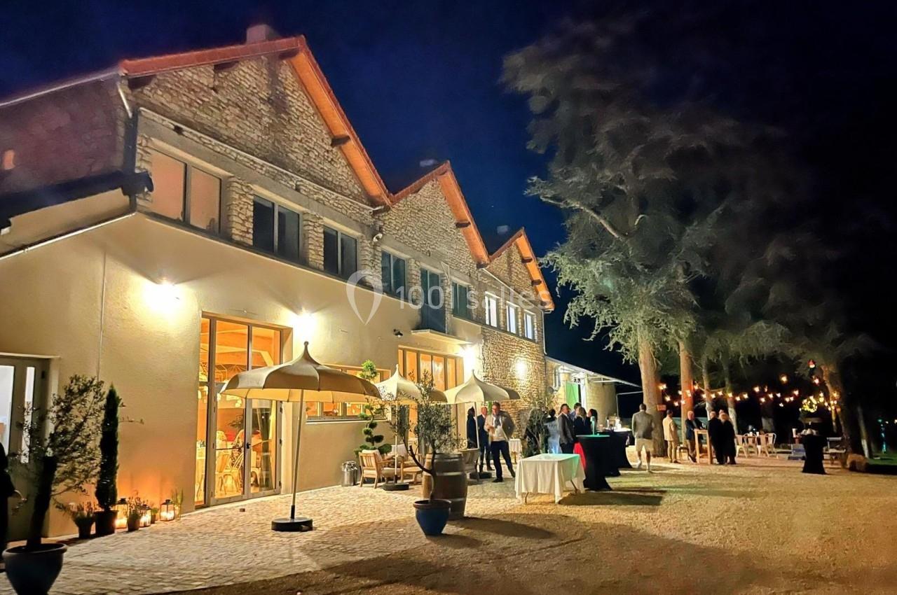 Façade éclairée d'un bâtiment en pierre de nuit, avec des invités rassemblés sur une terrasse aménagée.