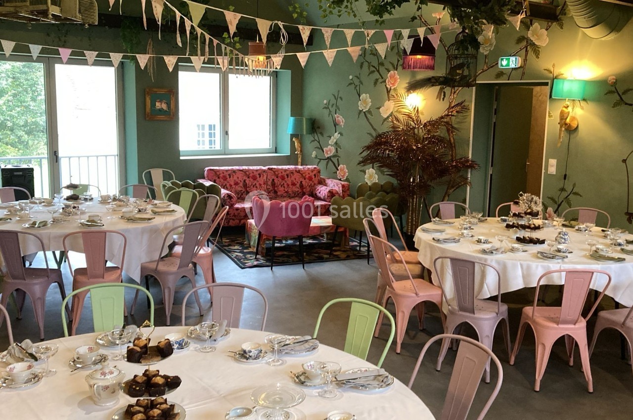 Salle décorée avec des tables rondes dressées pour un repas, chaises colorées et ambiance florale festive.