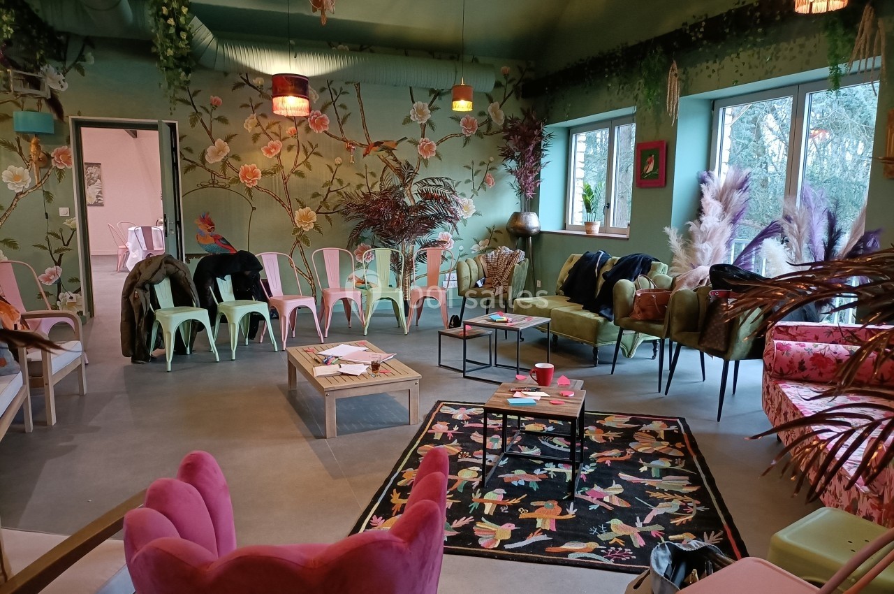 Salle colorée avec des chaises dépareillées, murs fleuris, plantes décoratives et lumière naturelle entrant par les fenêtres.