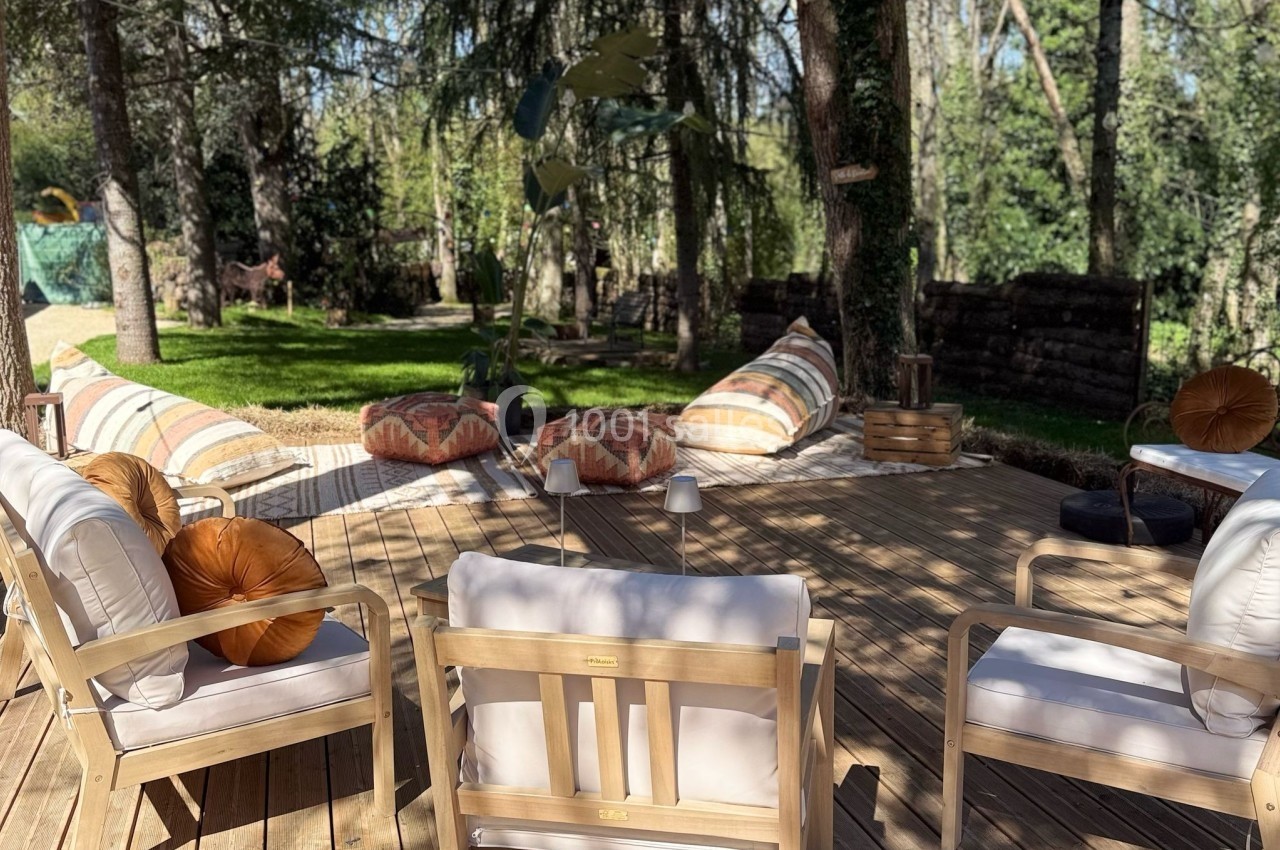 Terrasse en bois aménagée avec des fauteuils, coussins et poufs, entourée d'arbres dans un cadre naturel.
