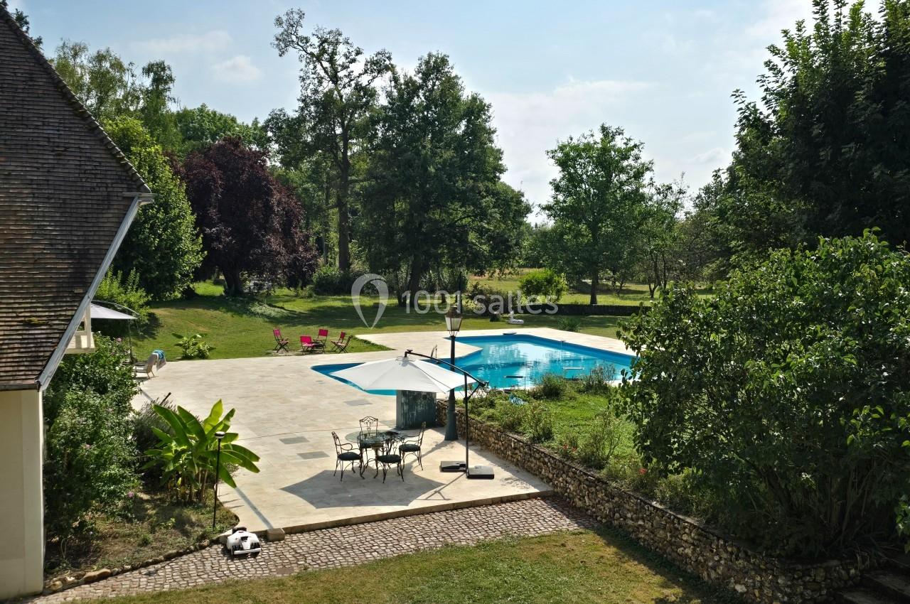 Terrasse avec parasol et mobilier de jardin près d'une piscine, entourée d'arbres et d'un grand espace vert.