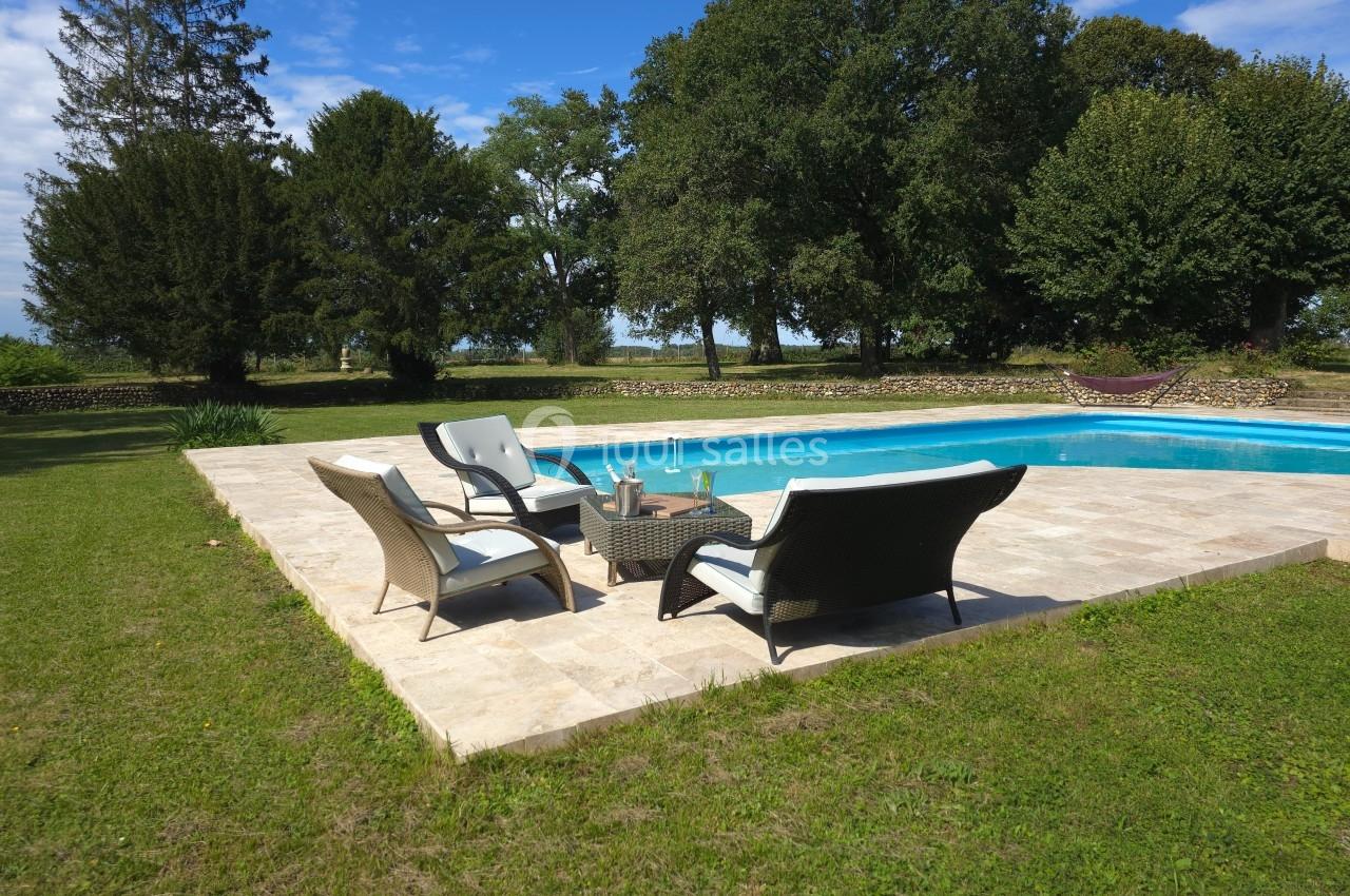 Mobilier d'extérieur autour d'une piscine sur une terrasse en pierre, entourée de pelouse et d'arbres.