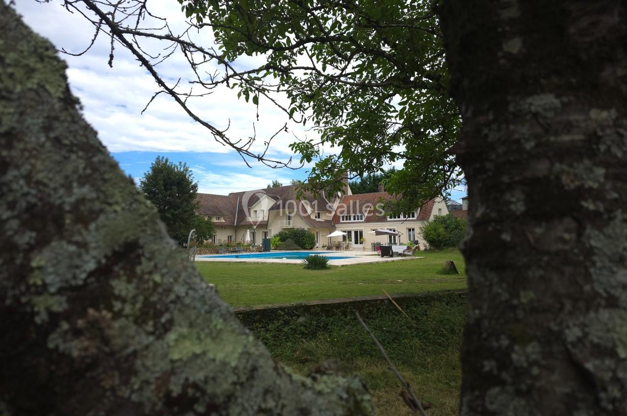 Vue d'une maison avec piscine, entourée d'un grand jardin, cadrée entre les branches d'un arbre au premier plan.