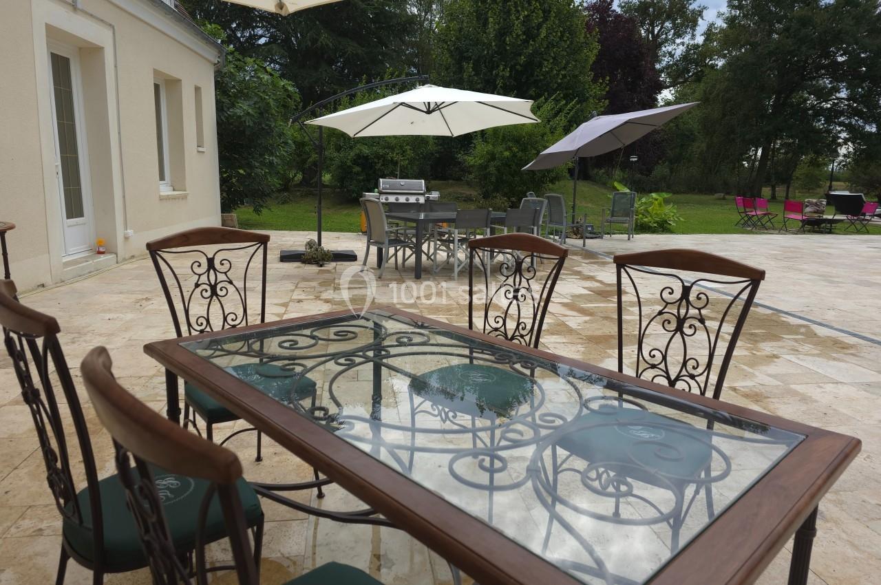 Table en verre avec chaises en fer forgé sur une terrasse en pierre, entourée de parasols et d'un jardin arboré.