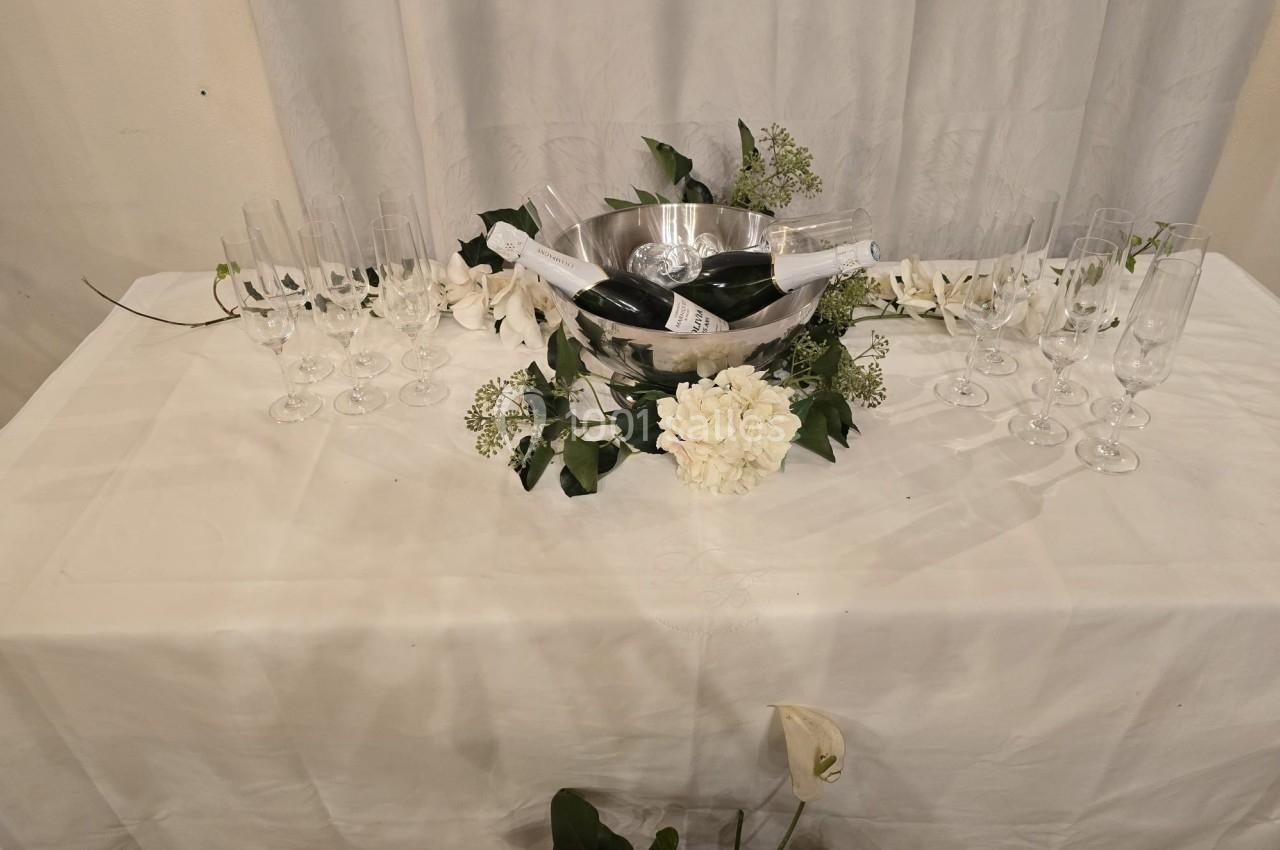 Seau en métal avec bouteilles de champagne entouré de fleurs blanches, posé sur une table avec des flûtes alignées.