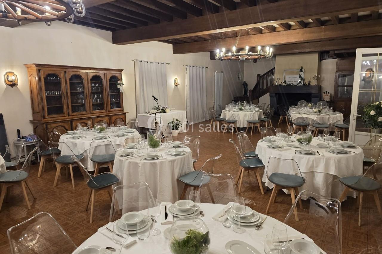 Salle de réception avec tables rondes dressées, chaises transparentes et décoration sobre dans un cadre rustique.