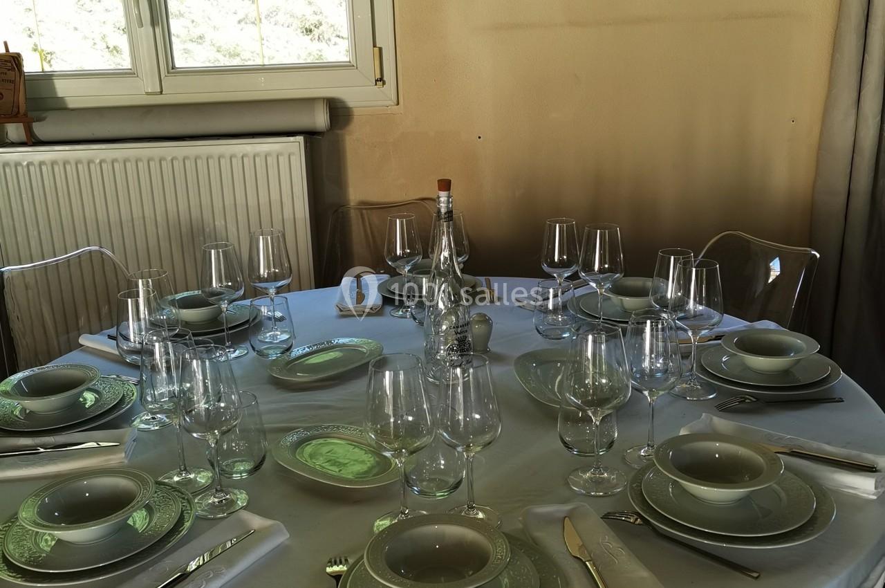 Table ronde dressée avec nappes blanches, assiettes, verres et couverts, près d'une fenêtre éclairant la pièce.