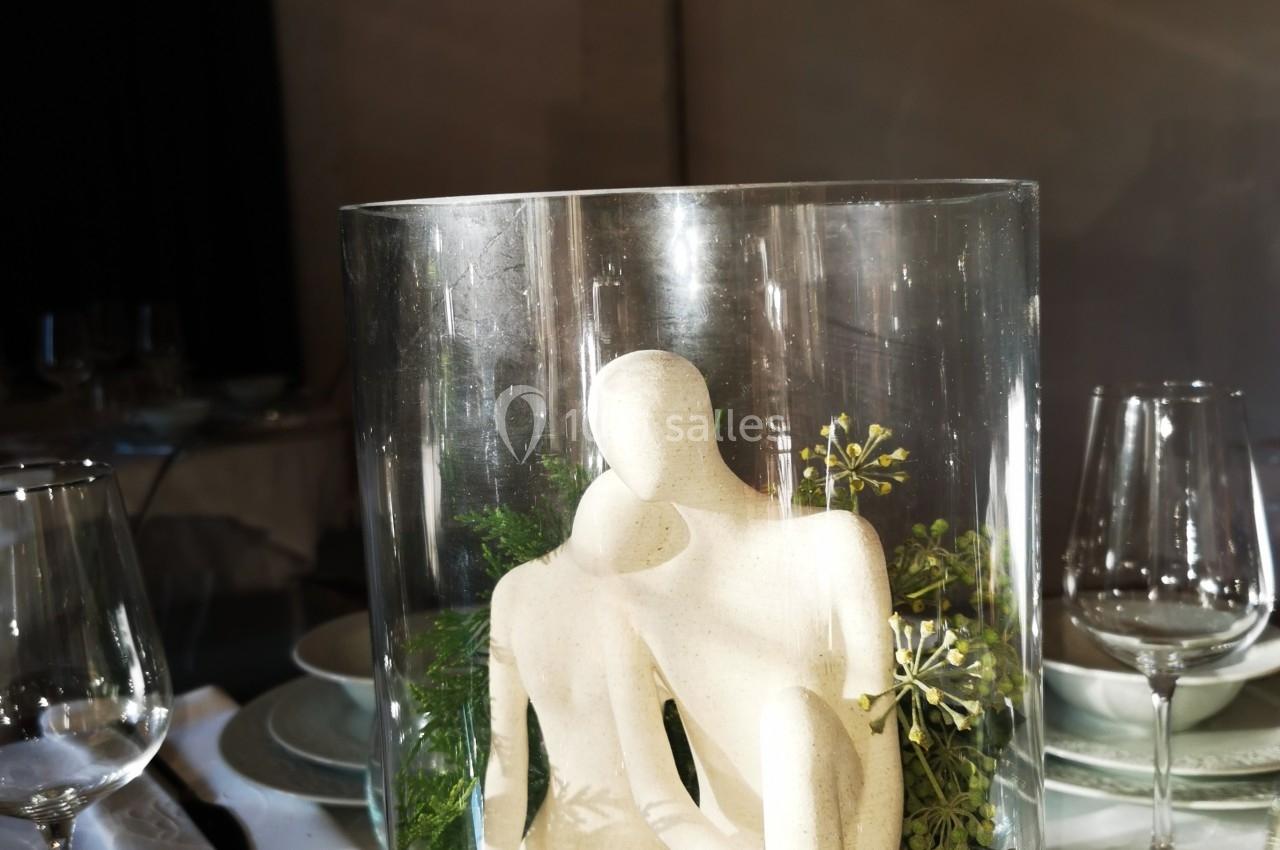 Deux sculptures abstraites blanches dans un vase en verre, entourées de feuillage, sur une table dressée.