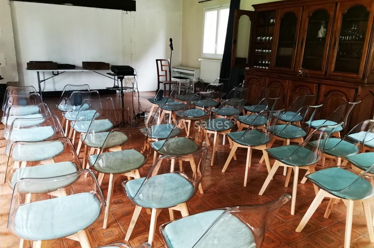 Salle aménagée avec des chaises transparentes et coussins bleus, face à un espace avec micro et tables.