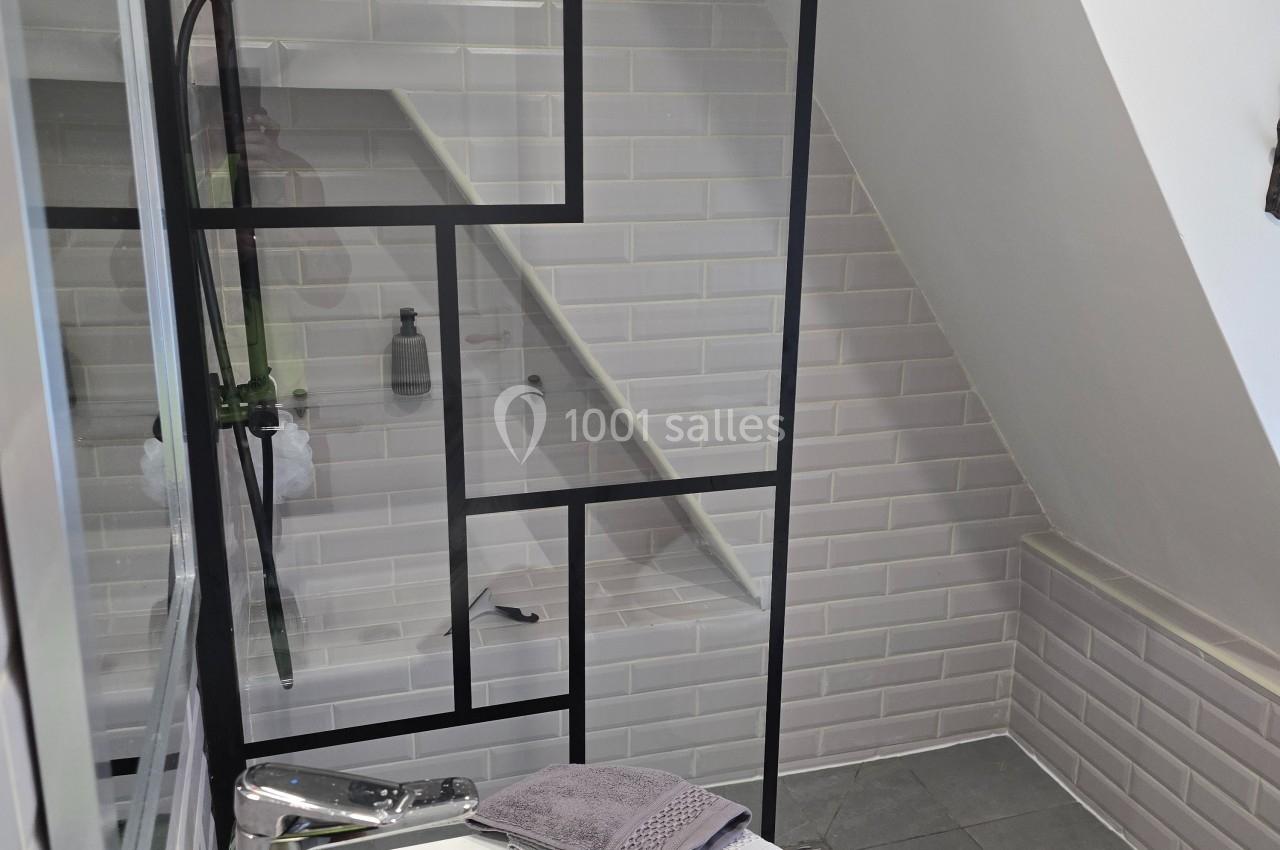 Douche moderne avec paroi vitrée à cadre noir, carrelage blanc et lavabo avec serviette grise dans une salle de bain…