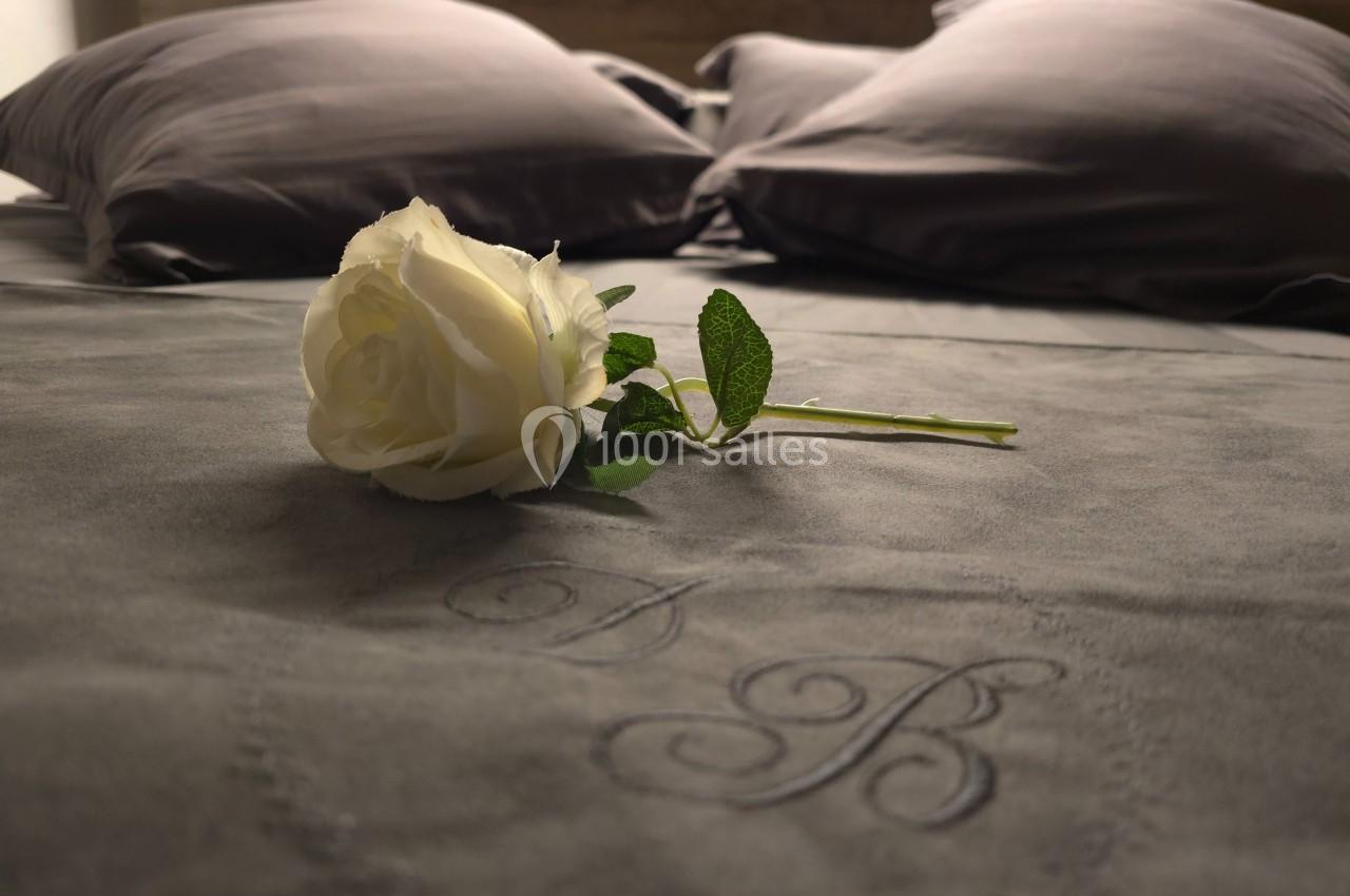 Une rose blanche posée sur un lit avec des draps gris et des initiales brodées sur la couverture.