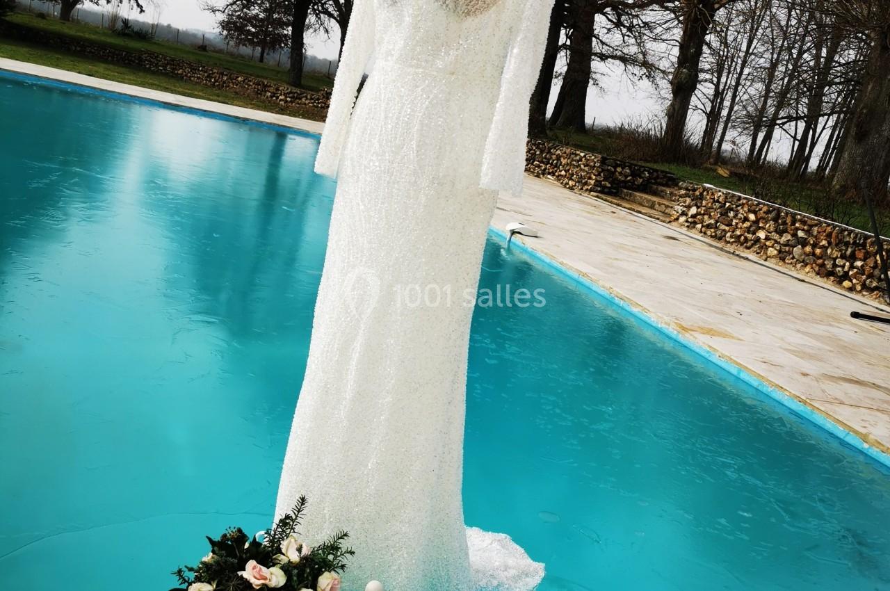 Robe de mariée blanche exposée sur un mannequin devant une piscine, accompagnée d'un bouquet de fleurs.