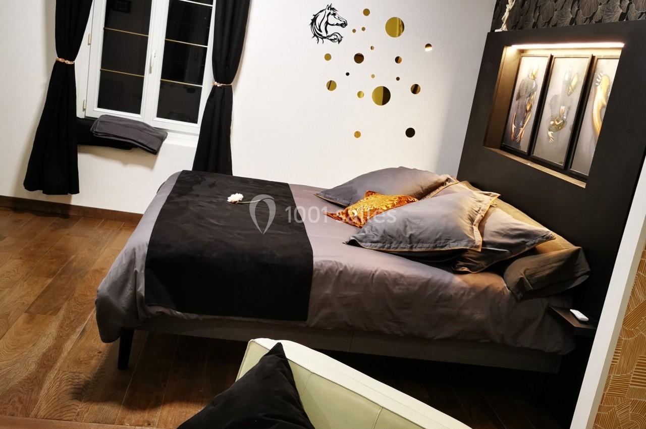 Chambre avec lit double, draps sombres, coussins colorés, fauteuil beige, fenêtre avec rideaux noirs et décoration murale.