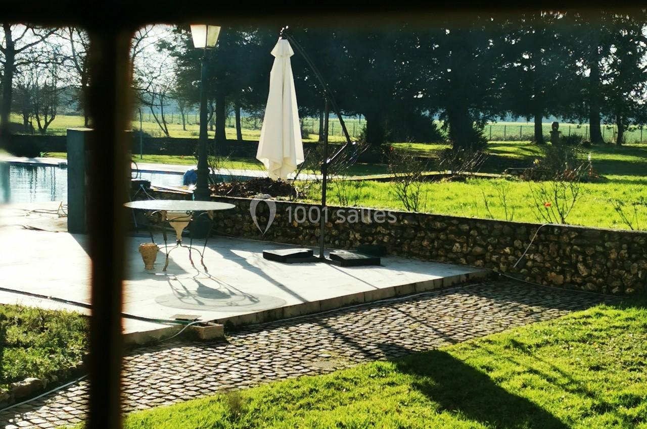 Vue depuis une fenêtre sur une terrasse ensoleillée avec parasol blanc, entourée de pelouse et d'arbres.